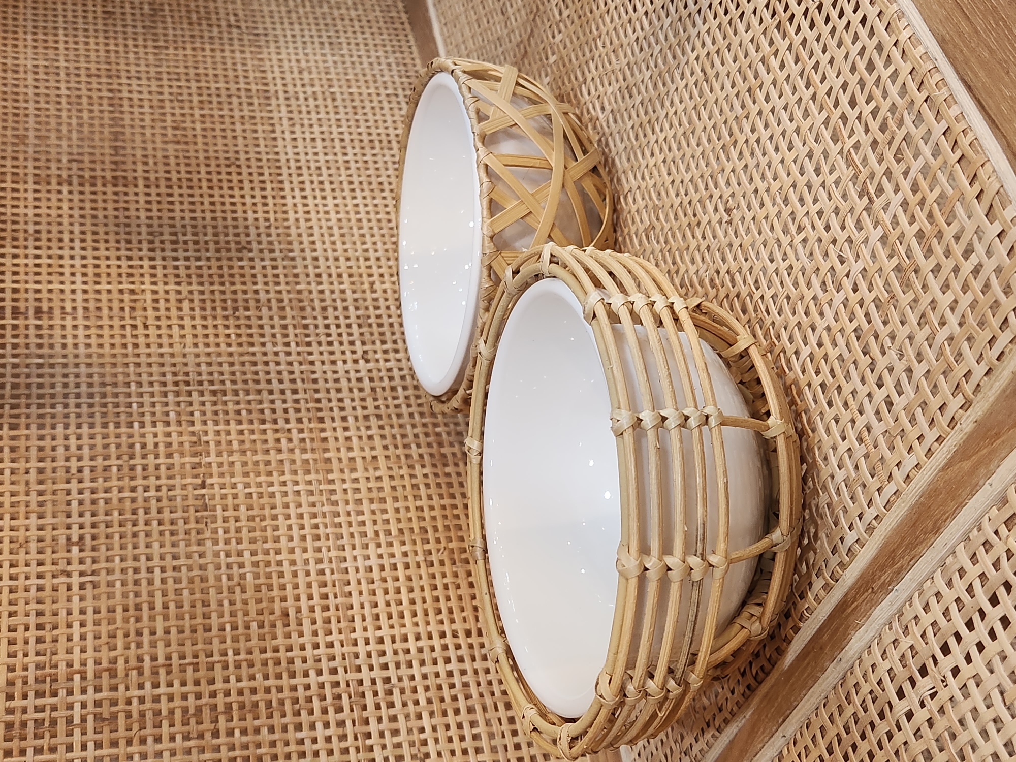 This is a medium shot, eye-level, outdoor photograph of two white ceramic bowls nestled in woven rattan baskets. The bowls are empty and facing the viewer at slight angles. The basket in the foreground is larger and closer to the camera, displaying horizontal bands of woven rattan. The basket in the background is smaller and slightly further away, featuring a criss-cross weave pattern. Both baskets are placed on a flat surface covered with a woven rattan mat, which extends to the edges of the frame. Behind the baskets, a wall or partition made of woven rattan creates a textured backdrop. The lighting is neutral, casting soft shadows. No people or animals are present. The scene is set on a shelf or table. The city of Maitland, United States of America, is not discernible from the image.