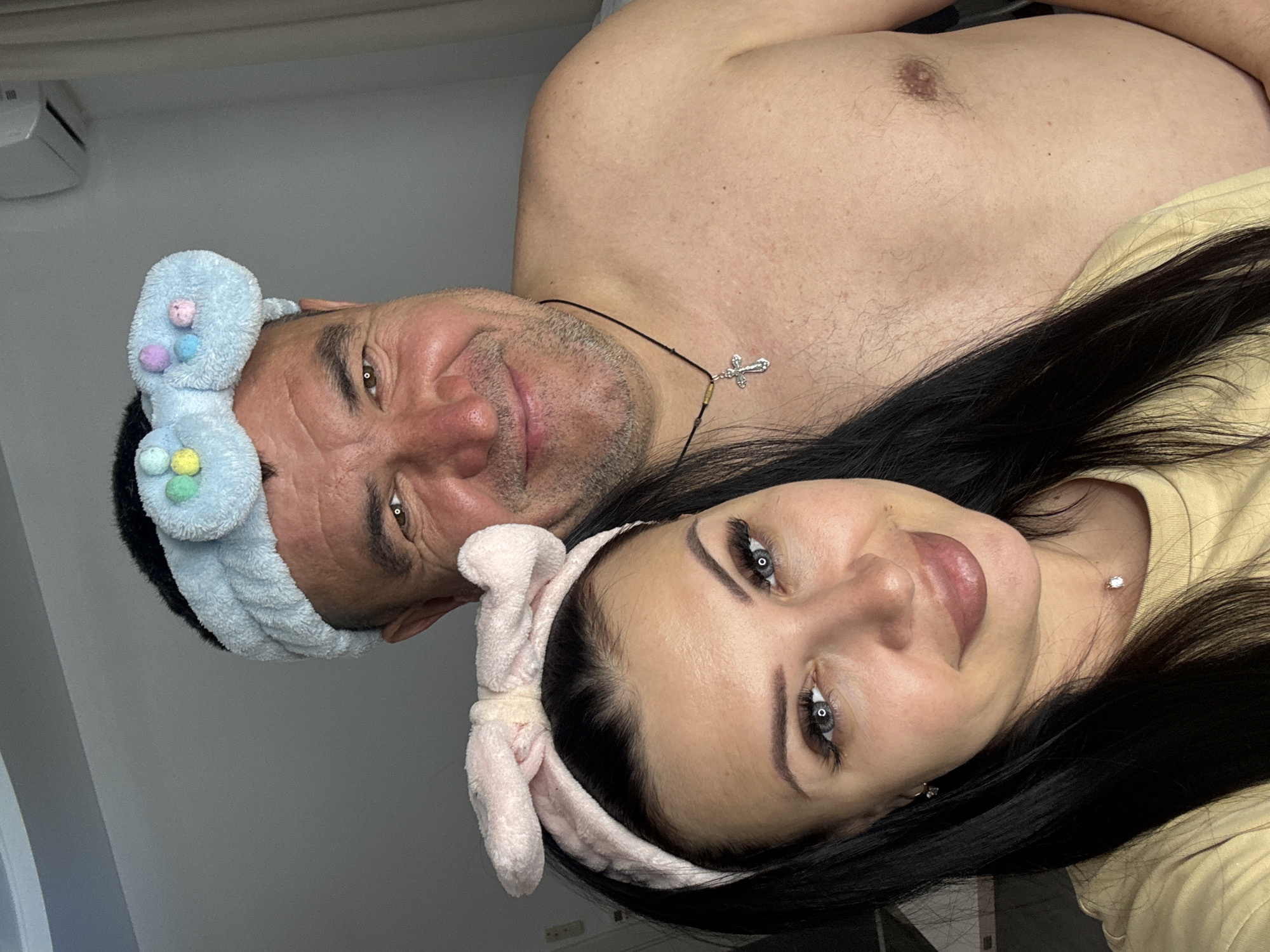 A selfie shows a bare-chested man with a blue headband beside a woman with long black hair, also wearing a headband. The man has a cross necklace. The setting appears to be indoors, possibly a home. The location is not discernable from the image.