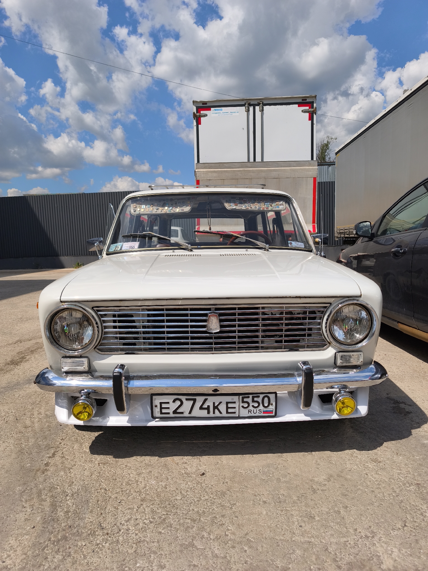 A frontal view captures a white vintage Lada (VAZ-2101 or similar model) parked outdoors in Molokovo, Russia, under a bright blue sky dotted with white cumulus clouds. The car features a prominent chrome grille with the Lada emblem, circular headlights, and a shiny chrome bumper with black bumper guards. Below the bumper, two yellow auxiliary fog lights are attached. The license plate, "E274KE 550 RUS," clearly identifies it as a Russian vehicle from the Omsk region (550). The car's windshield is adorned with numerous small decorative ornaments along the top edge, and a document is visible on the dashboard. A silver roof rack is also present.
To the right of the Lada, the front section of another dark-colored car is partially visible. In the background, behind the white car, a large white box truck or trailer is parked, displaying red markings on its rear doors and partial Russian text that reads "ПРОДАЖА, СЕРВИС, TRADE-IN" and "АВТОЦЕНТР ГАЗ" (Sale, Service, Trade-in, GAZ Auto Center). Further back, a dark grey corrugated metal fence defines the perimeter of what appears to be an industrial or commercial lot. The ground is a light-colored concrete or asphalt. The clear sky and distinct shadows indicate a sunny daytime setting. No people are visible.