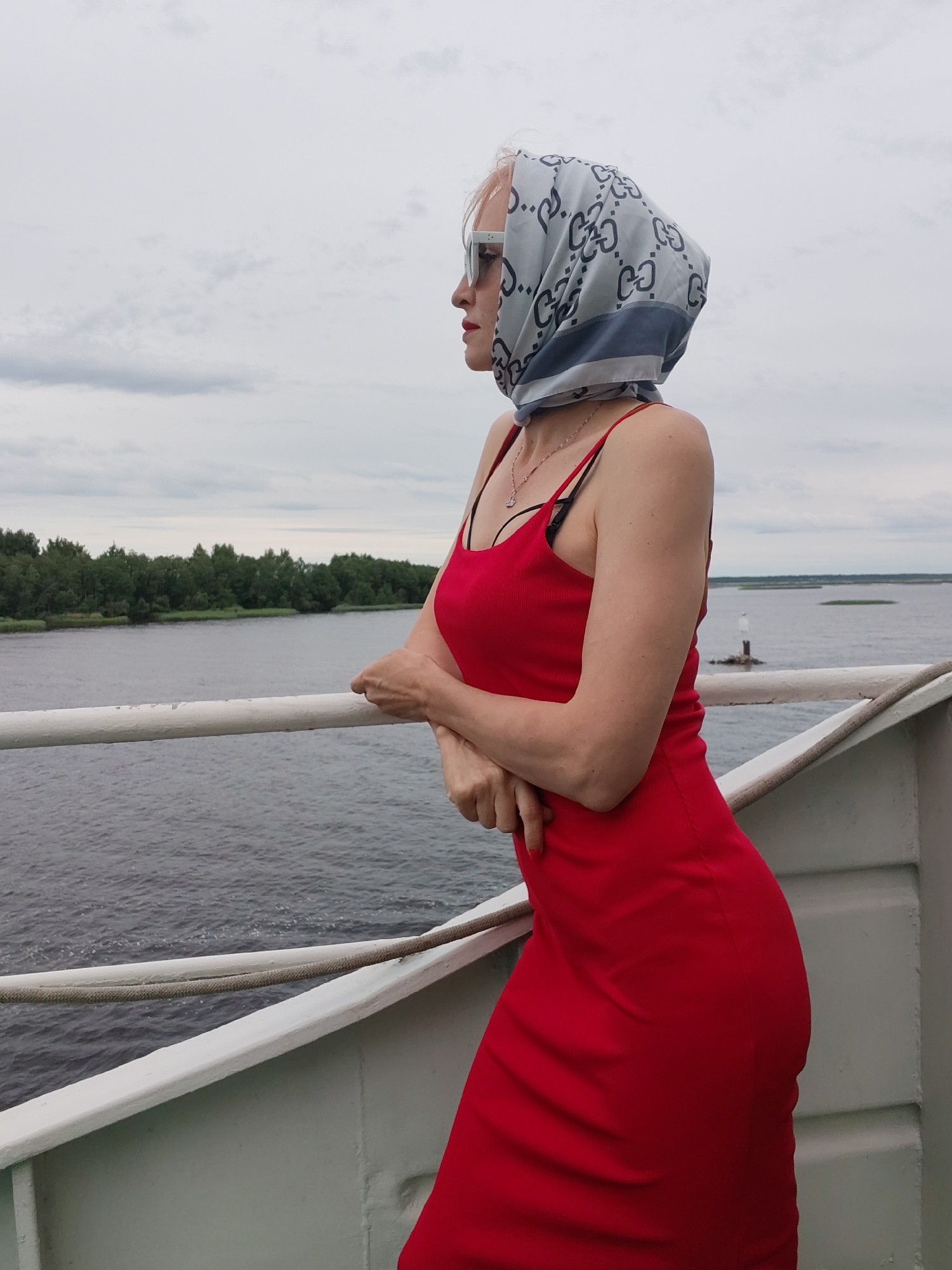 A woman stands on the deck of a ship, looking out at a wide body of water. She is wearing a red, form-fitting dress and a light blue headscarf with a repeating gray pattern. She has on white sunglasses and a delicate silver necklace. Her arms are crossed in front of her as she leans against the ship's railing. The water is choppy and gray, suggesting a cloudy day. In the distance, a line of trees forms the shoreline, and a small lighthouse or buoy stands in the water. The sky is overcast with thick, gray clouds. The overall mood is contemplative, with the woman seemingly lost in thought as she gazes at the horizon. The setting appears to be a river or a large lake in Bereg, Russia. The time of day is difficult to determine due to the cloudy conditions, but it appears to be daytime.