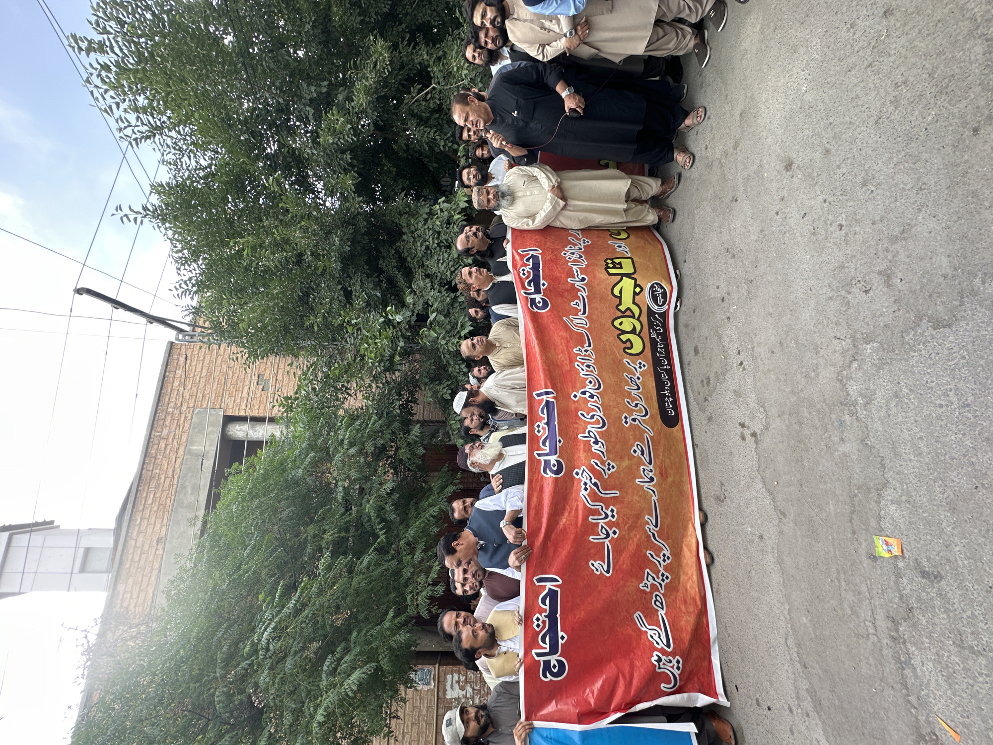 A group of men are holding a banner in Quetta City Tehsil, Pakistan. The banner is in Urdu and appears to be related to a protest regarding loans and the removal of smart locks. The group is standing on a street in front of a brick building with a tree.