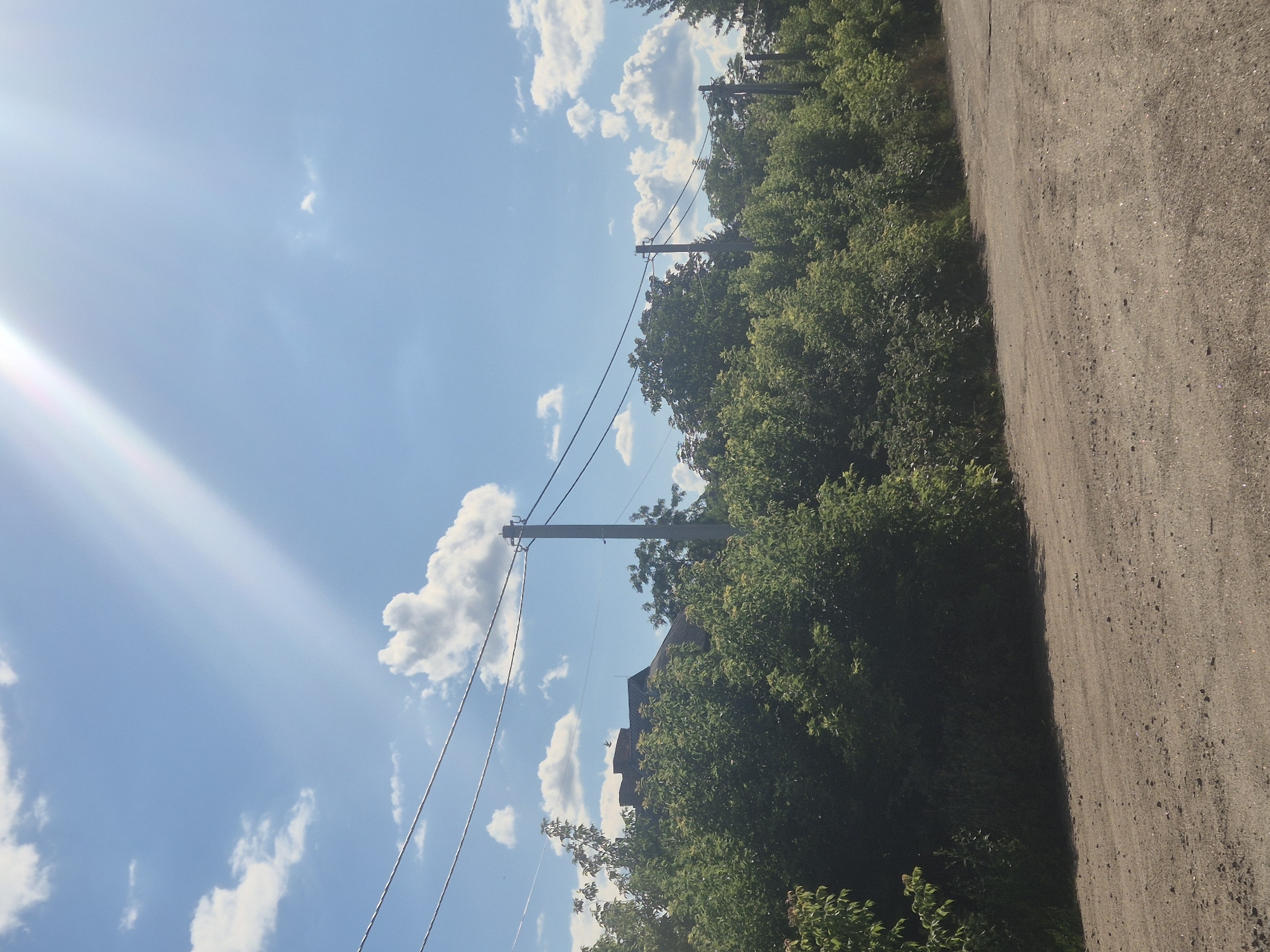 In Sozonivka, Ukraine, a rural landscape unfolds under a bright, sunny sky with scattered white clouds. A dirt road curves through the foreground, bordered by a dense line of lush green trees and bushes. In the background, telephone wires stretch across the frame, supported by utility poles. A building with a dark, sloped roof and a brick chimney is partially visible through the foliage. The sun casts a strong, diagonal ray across the sky, creating a lens flare effect. The scene appears peaceful and depicts a typical rural setting.