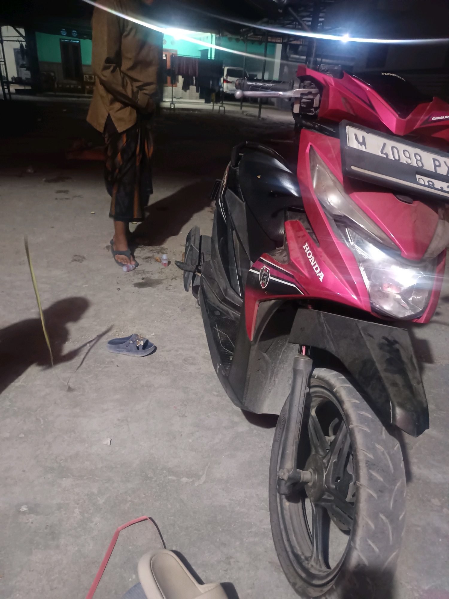 A medium shot captures a pink Honda scooter parked on a concrete surface at night. The scooter's front is angled towards the viewer, with its headlight illuminated. A license plate reading "M 4098 PX" is visible. To the left of the scooter, a person wearing a brown jacket and a patterned sarong stands with their feet in a pair of dark blue sandals. Another pair of dark blue sandals lies on the ground near the person's feet. A thin yellow stick casts a long shadow on the concrete. In the background, a building with green walls and clothes hanging on a line is dimly lit. The scene appears to be outdoors, possibly in a residential or commercial area in Gunung Sekar, Indonesia, given the context.