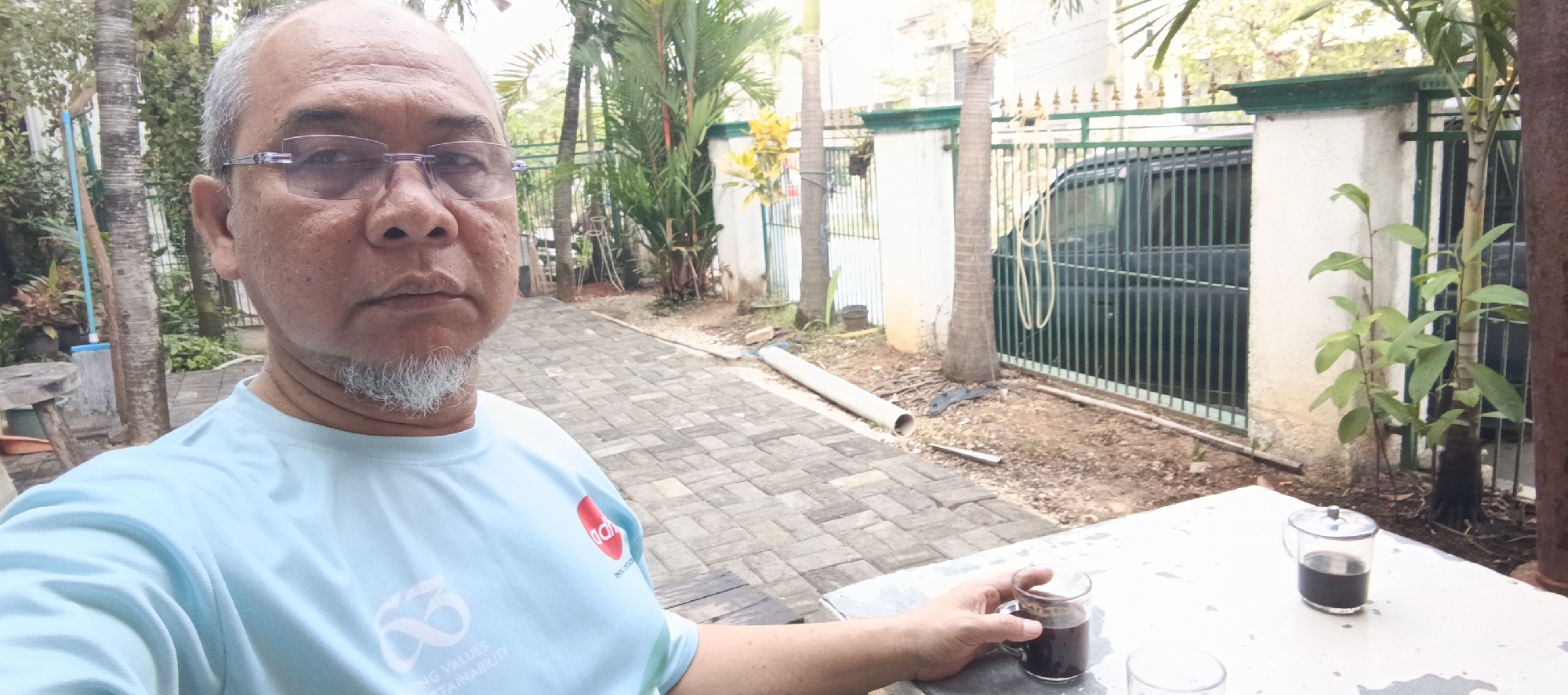 A middle-aged man with a goatee and glasses is taking a selfie outdoors. He is wearing a light blue t-shirt with a logo on the left side of his chest. The man is standing on a paved walkway, with a table to his right. On the table are two clear glasses, one containing a dark liquid, presumably coffee. Behind the man, there is a fence, a dark-colored vehicle, a concrete pillar, and several types of plants, including palm trees. The environment suggests a residential area or a park, possibly in Sukaresmi, Indonesia, given the style of fencing and vegetation.