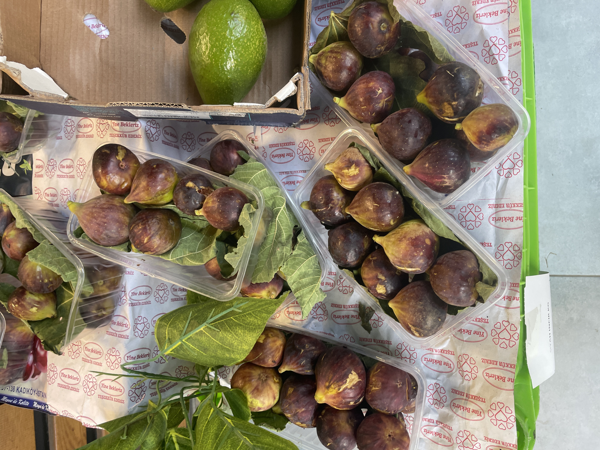 The image shows a close-up, overhead view of a market stall displaying fresh figs and avocados. The figs are arranged in clear plastic containers, with some leaves from the fig trees visible. The containers are placed on white paper with a repeating pattern of hearts and text in Turkish, which reads "Yine Bekleriz" (We await you again) and "Teşekkür Ederiz" (Thank you). The background includes cardboard boxes, suggesting a retail environment. One of the containers has a label at the bottom with Turkish text that includes "SİYAH İNCİR KG" (Black Fig KG), indicating the product being sold. The stall is located in Kadıköy, Turkey, as indicated by the text on a paper visible in the upper left corner of the image, which reads "138 KADIKÖY İSTANBUL". There are no people visible in the image, and the focus is entirely on the produce and the stall's presentation.