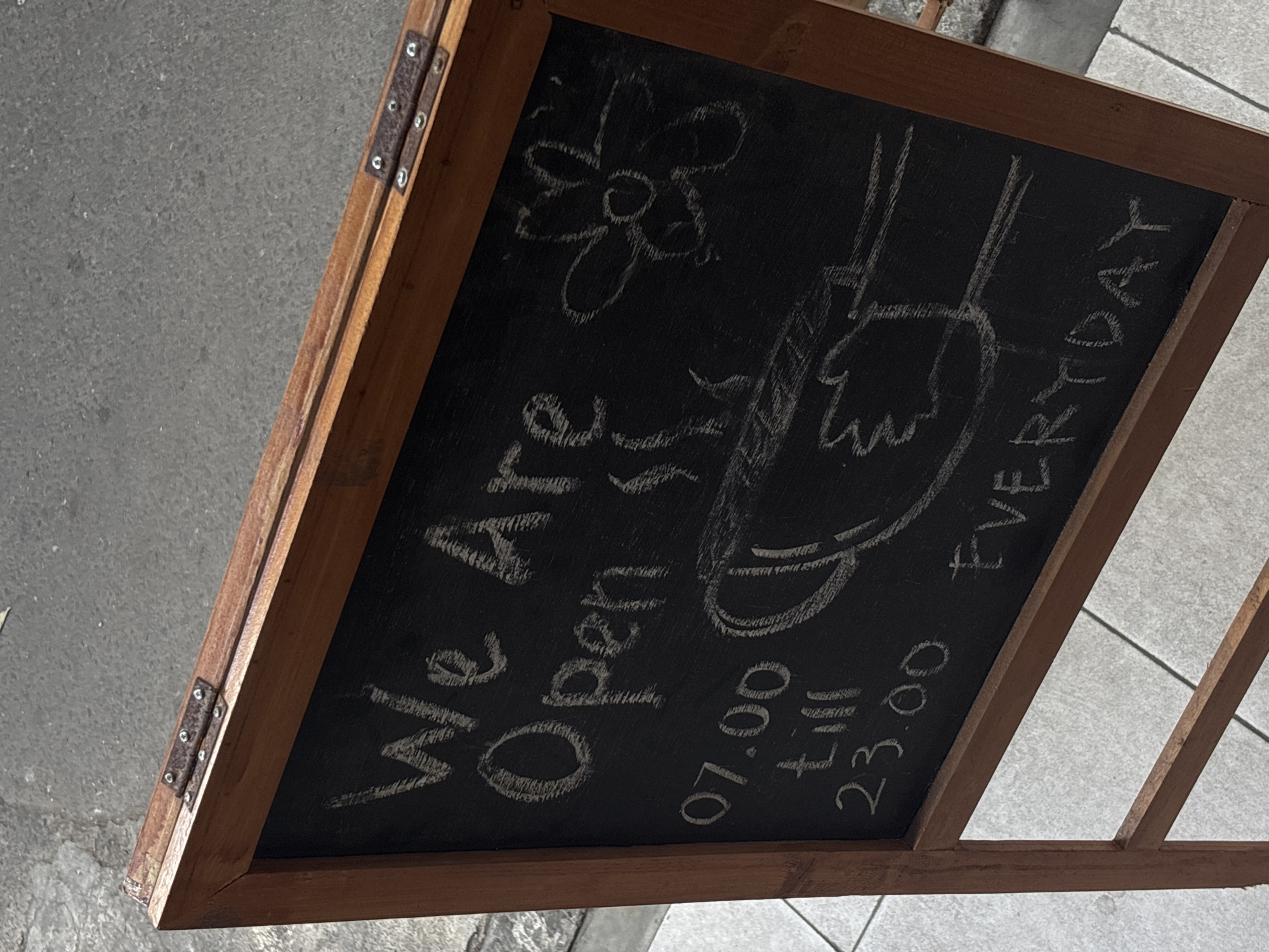 A wooden A-frame chalkboard sign stands on a paved walkway. The sign is oriented at an angle to the viewer, with the top of the frame closer than the bottom. The chalkboard surface is black and has white chalk writing and drawings on it. The writing says "We Are Open" in large letters, followed by "07.00 till 23.00 EVERYDAY". There are also chalk drawings of a flower and a hand holding a cup with steam rising from it. The wooden frame has hinges on the top edge, suggesting it is foldable. The background shows a rough, gray paved surface that appears to be a sidewalk or street, and a lighter-colored tiled surface on the left. The scene suggests a business establishment that is open for customers. The context provided indicates the location is in Semarang, Indonesia, but this cannot be visually confirmed from the image itself.