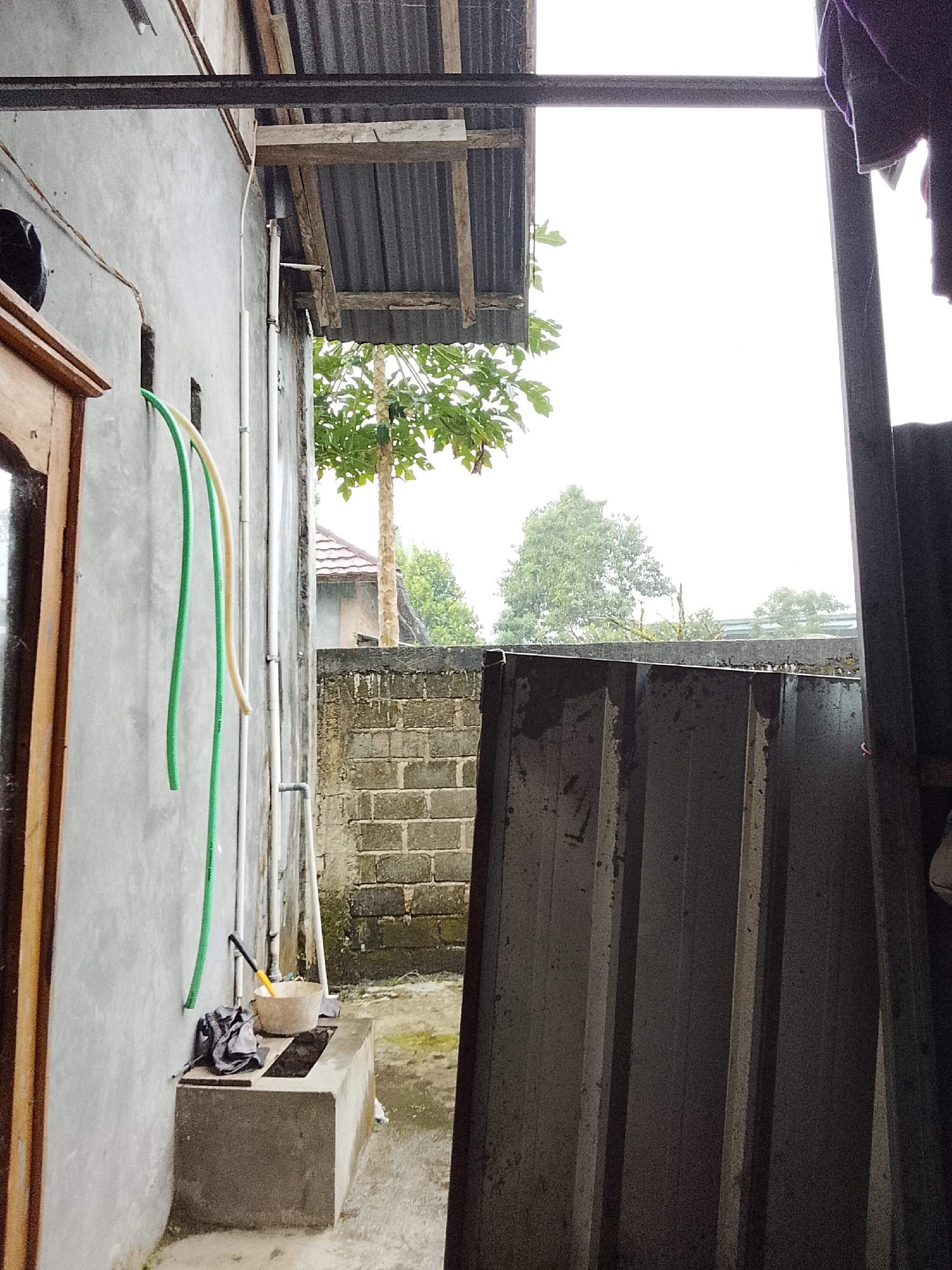 The image depicts an outdoor scene, likely a residential backyard or utility area, under an overcast sky. On the left, a grey concrete wall is visible, with a wooden doorframe on the far left. Two colorful hoses, one green and one light yellow, hang vertically from the wall. Below them, a grey cloth is placed on a concrete structure, which appears to be a small sink or water basin with a yellow-handled tool inside a beige bucket. A concrete partition wall made of bricks is situated behind this basin, with a large, dark grey corrugated metal sheet leaning against it in the foreground on the right. Beyond the brick wall, trees and foliage are visible, indicating a vegetated environment. The overall impression is one of a functional, perhaps somewhat utilitarian, outdoor space, possibly experiencing damp weather. The context of "Padang Dalom, Indonesia" suggests this location is in a tropical region.