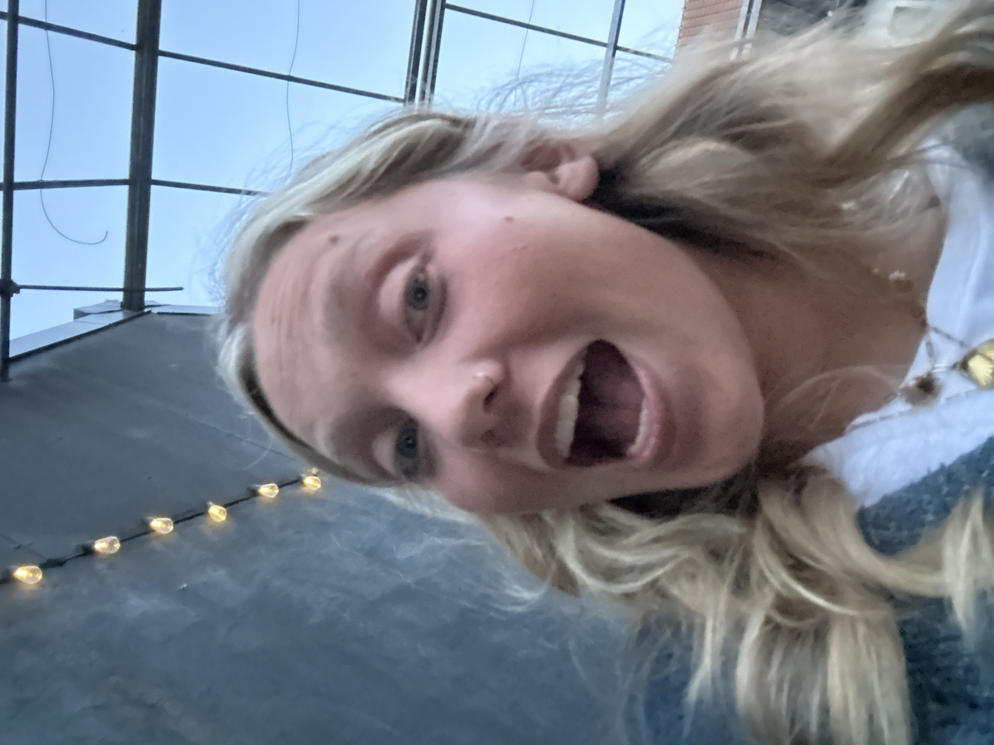 A young woman with blonde hair is captured from an extreme low-angle perspective, looking directly down at the camera. Her mouth is open wide in an expression that conveys excitement, surprise, or laughter, and her light-colored eyes are visible. She is wearing a white top and a blue or grey knitted cardigan, along with a necklace featuring a golden, possibly floral, charm.
The background reveals a clear, light blue sky intersected by dark, geometric structural beams, which could be part of a skylight, a glass roof, or an architectural framework. In the lower left, a string of warm, yellow fairy lights illuminates a dark, flat surface, possibly a floor or roof. The lighting suggests it is daytime or early evening with clear weather. The image evokes a playful and energetic mood. No text is visible. The provided location context is Hem, France.
