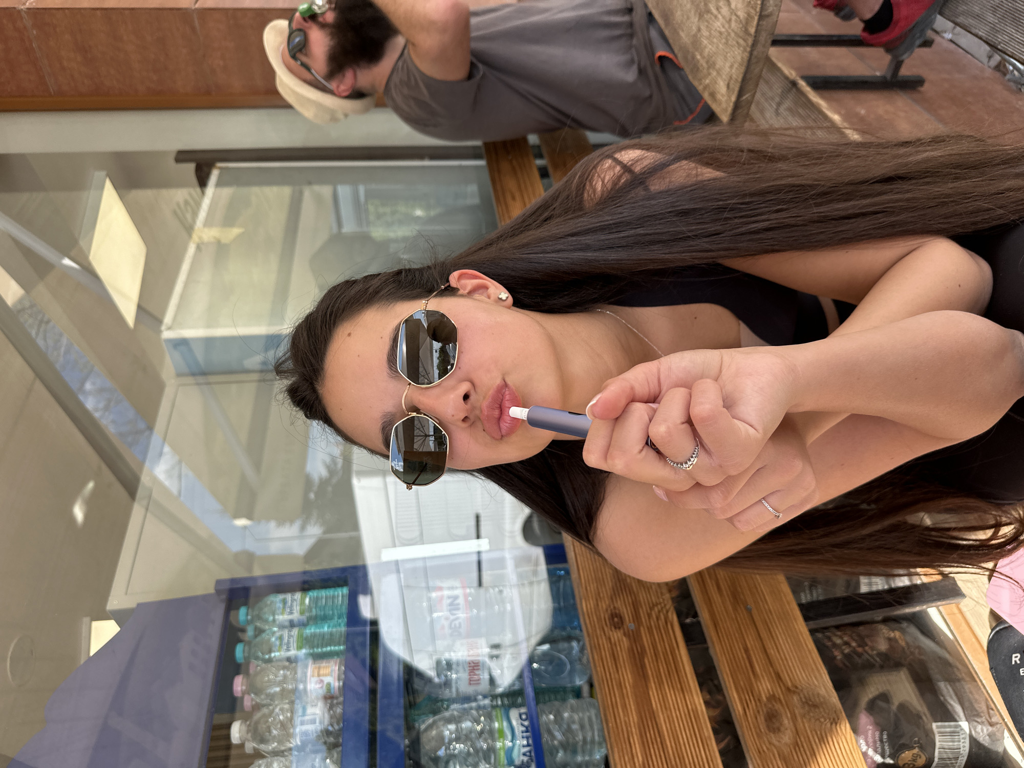 A woman with long, dark hair and sunglasses is sitting on a wooden bench, holding a blue IQOS device to her lips. Behind her is a glass window showcasing a refrigerated display case filled with bottles of water. The bottles feature Bulgarian labels, suggesting the location is in Bulgaria. A man with a beard and sunglasses is visible in the background, reclining on a bench near the woman. The presence of Cyrillic script on the water bottles indicates the scene is likely in a city within Bulgaria, possibly Plovdiv. The presence of water bottles and the bench implies a semi-public space like a cafe or shop terrace.