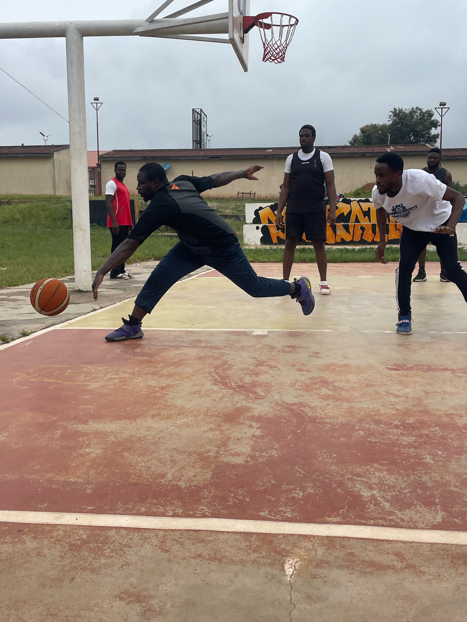 This is a medium-shot, eye-level photograph taken on an outdoor basketball court in Gura Top, Nigeria. The court's surface is a worn, reddish-brown and yellow concrete with white boundary lines. In the background, there are beige-colored buildings with red roofs, a metal basketball hoop and backboard, and some trees. The sky is overcast.
In the foreground, a man in a black and grey basketball jersey and dark pants is in motion, dribbling a basketball. He is leaning forward, with his left leg extended and his right leg bent. The basketball is in mid-air, to the left of his body, suggesting he has just released it. His right arm is extended towards the ball, with his hand open.
Behind him, to the right, another man in a white t-shirt with a graphic print and black athletic pants is crouched in a defensive stance, facing the dribbling player. Further back, a man in a black sleeveless jersey and black shorts stands observing the game. To the far left, a man in a red basketball jersey and dark pants is partially visible, looking towards the action. The overall atmosphere suggests a casual game of basketball is taking place.