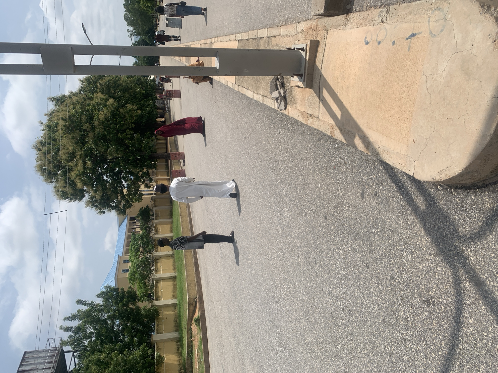 The image depicts a street scene in Jalingo, Nigeria. Several people are visible on the asphalt road, which is lined with a concrete sidewalk. The time of day appears to be daytime, with clear skies and sunlight casting shadows.
In the foreground, a person in a white garment stands facing away from the camera. Further up the road, another individual in dark clothing and a patterned shirt is also standing. In the middle ground, a person in a maroon abaya is standing, and behind them, another person in light brown clothing is visible. In the background, more people are walking on the sidewalk.
A large green tree with blooming flowers is prominent on the left side of the image, behind a concrete pillar and some metal poles. A building with a blue roof and yellow walls is visible in the distance. The concrete sidewalk has some markings, including what appear to be numbers or symbols. The overall atmosphere seems to be one of everyday activity under a bright sky.