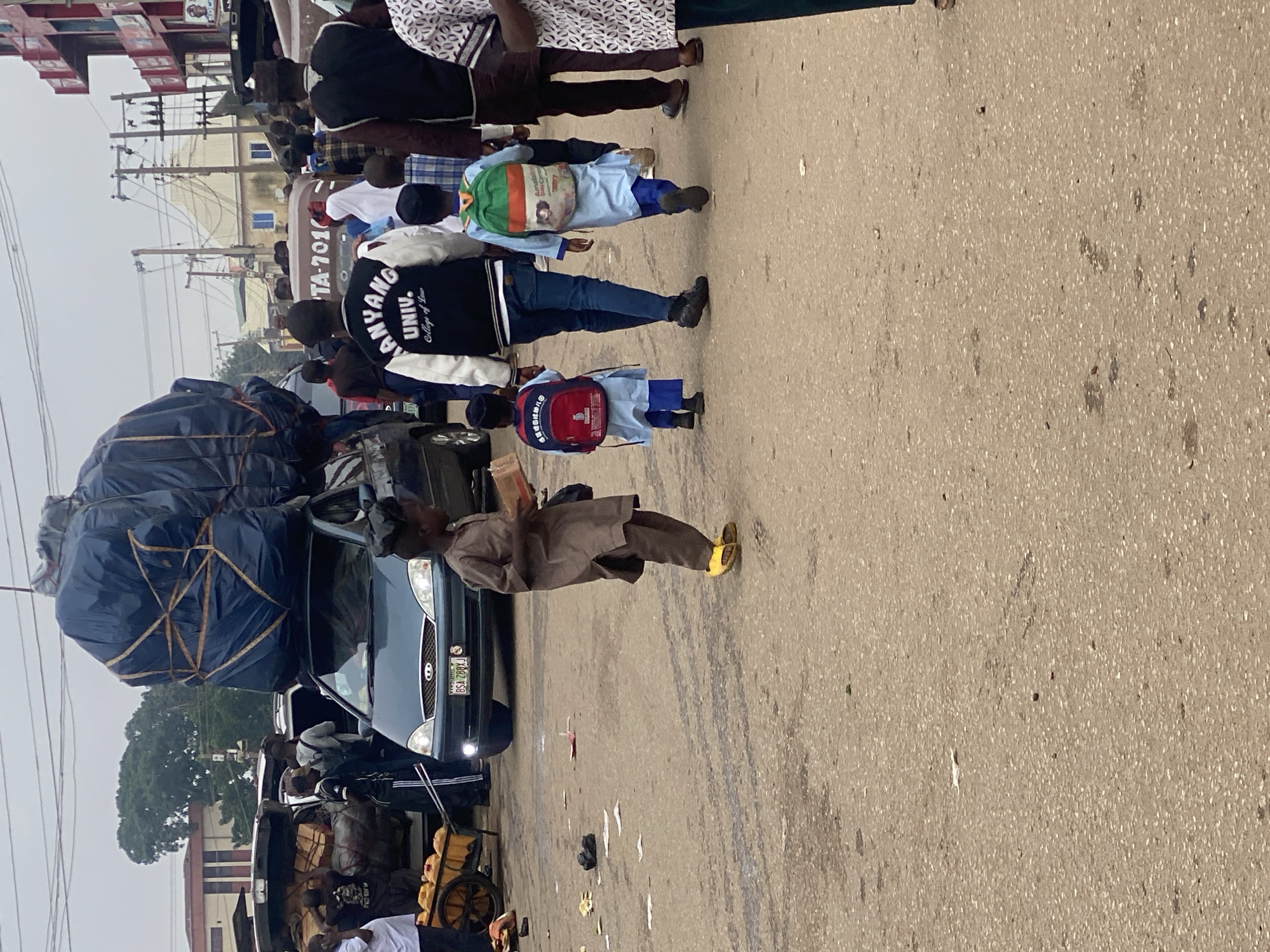In Jos, Nigeria, a busy street scene unfolds with a large vehicle piled high with dark blue tarp-covered goods. A young boy in a brown tunic and yellow sandals walks away from the camera, holding a small brown object. Behind him, two children in light blue tunics and matching caps walk with backpacks, one red and one green and orange. An adult in a black and white jacket with "ANYANG UNIV. College of Law" on the back walks ahead of them. Other people and vehicles are visible in the background, including a cart with yellow containers. The ground is a dusty, asphalt surface with scattered debris.