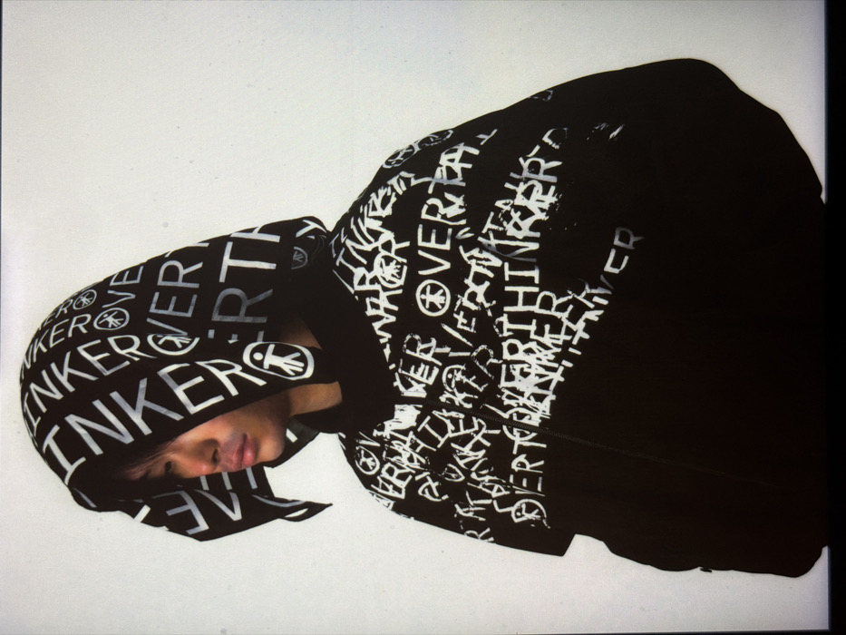 The image displays a head and torso shot of an individual, appearing male, with East Asian features. The person is positioned facing left, with the head slightly angled down, and is wearing a black hooded garment. The garment, which appears to be a jacket or cape, is extensively patterned with white, graffiti-style text and symbols. Prominently visible text includes repetitions of "OVERTHINKER," often overlapping. Repeated symbols include a stylized stick figure with a circular head and body, and a peace sign. The hood is pulled up, obscuring much of the hair. The garment features a zipper down the front, visible on the chest. The background is a plain, untextured white or off-white surface, characteristic of a studio environment. No contextual elements are present in the image that indicate a specific location in the city of London, United Kingdom.