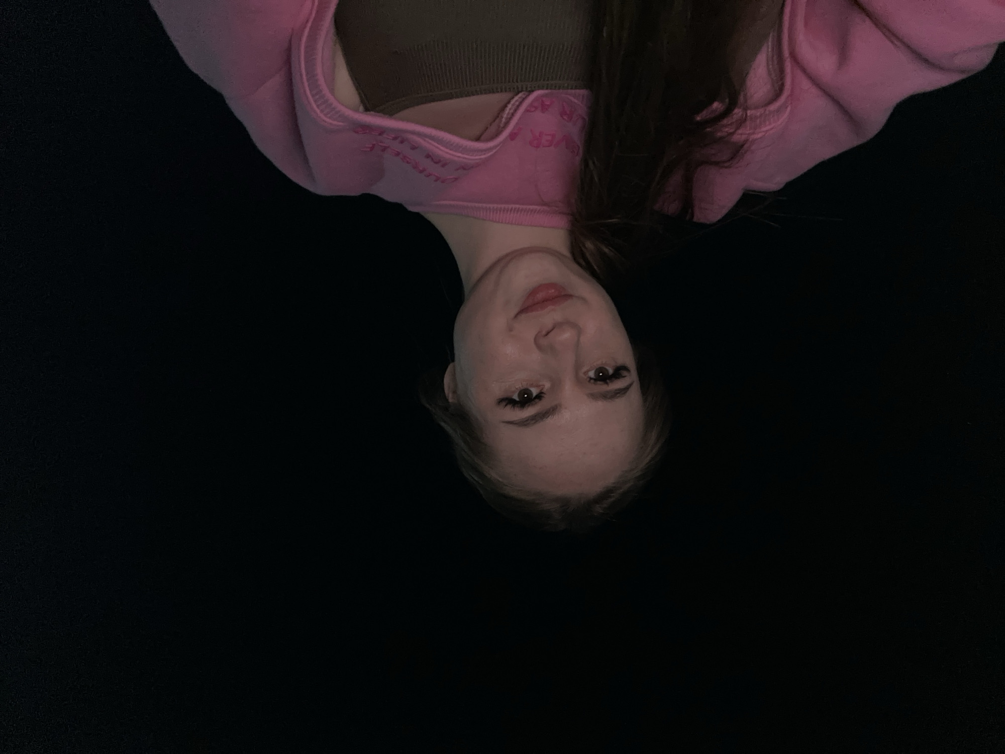 An inverted close-up portrait features a young woman against a uniformly dark, likely black, background. Her head and upper torso are visible, with her face oriented towards the bottom of the frame. She has fair skin, dark eyes enhanced with visible eyelashes or mascara, and her lips are slightly parted in a soft, neutral expression or subtle pout. Her dark hair frames her face and falls around her shoulders. She is wearing a light pink hooded sweatshirt or top, with ribbed cuffs and hem, over a dark olive green or brown inner garment, possibly a tank top or sports bra. Inverted text is visible along the collar area of the pink top, with partially discernible words such as "YOURSELF IN" and "EVER" printed in a lighter shade of pink or white. The lighting appears to come from above, illuminating her face and the upper part of her garments, while the rest of the scene remains in deep shadow, providing no cues about the location or time of day. She appears to be posing for a photograph.