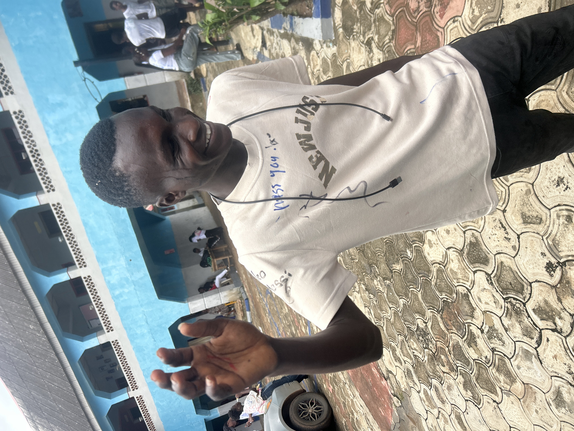 A young Black male, wearing a light-colored t-shirt with text and what appear to be blue markings, and black pants, smiles broadly while waving with his left hand. He has short, dark hair and is wearing a black cord around his neck. The background features a multi-story light blue building with balconies and white railings, typical of educational institutions in the area. The ground is paved with interlocking stone tiles. In the middle ground, other individuals, some wearing white shirts, are visible, suggesting a social gathering or event. A silver car is parked to the left. The overall environment appears to be an outdoor courtyard of a school or community center in Ikorodu, Nigeria.