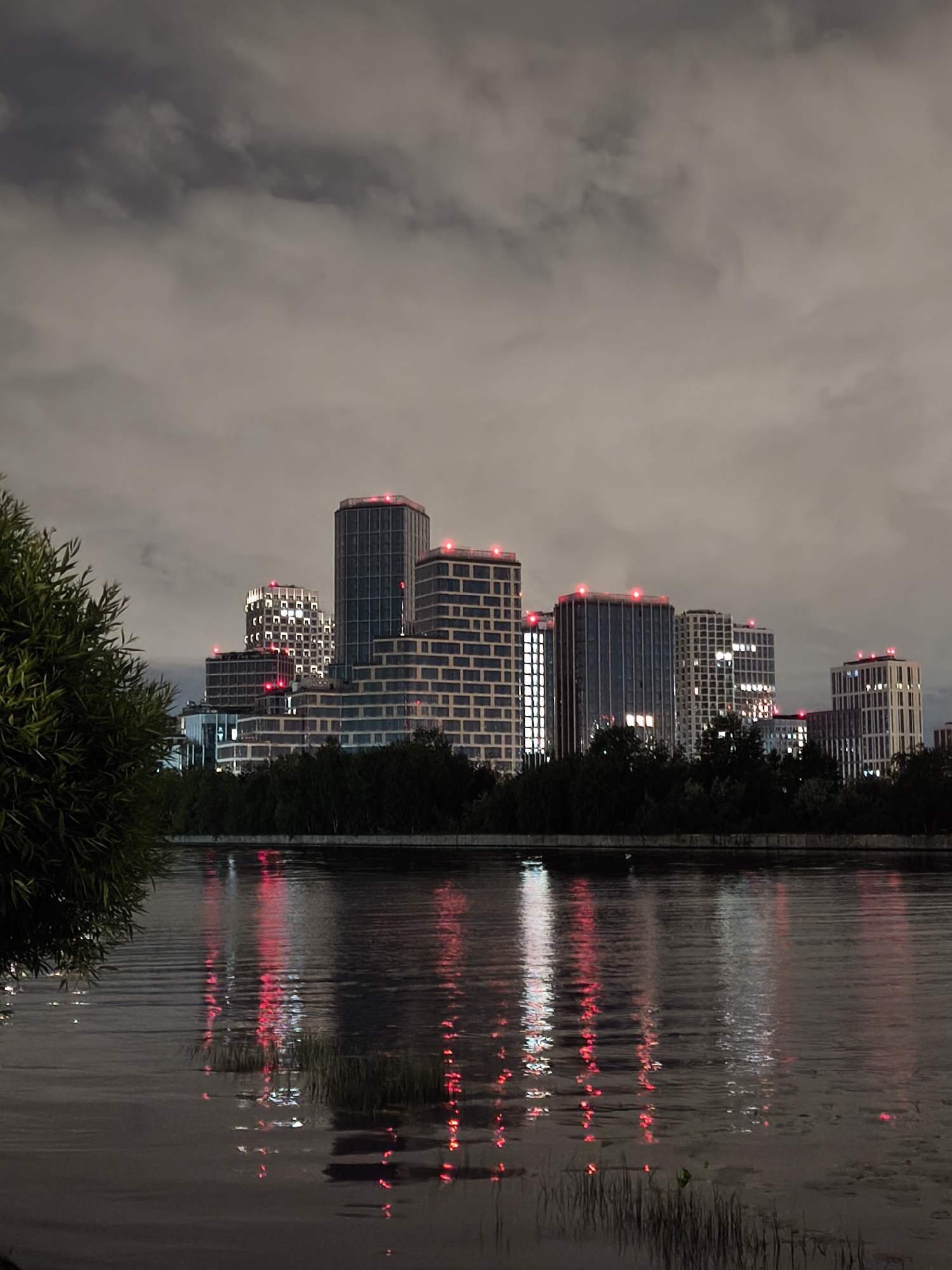 The image depicts a cityscape at night, likely in Moscow, Russia, due to the architectural style of the buildings. Several modern high-rise buildings are visible, illuminated with internal lighting and red lights atop each structure. These buildings are situated behind a line of trees, which obscure their lower portions. In the foreground, a body of water reflects the building lights as well as the red lights in the reflection on the water. The sky above is cloudy, contributing to the overall muted lighting of the scene. A portion of a tree is visible on the far left of the frame. The scene appears calm and still.