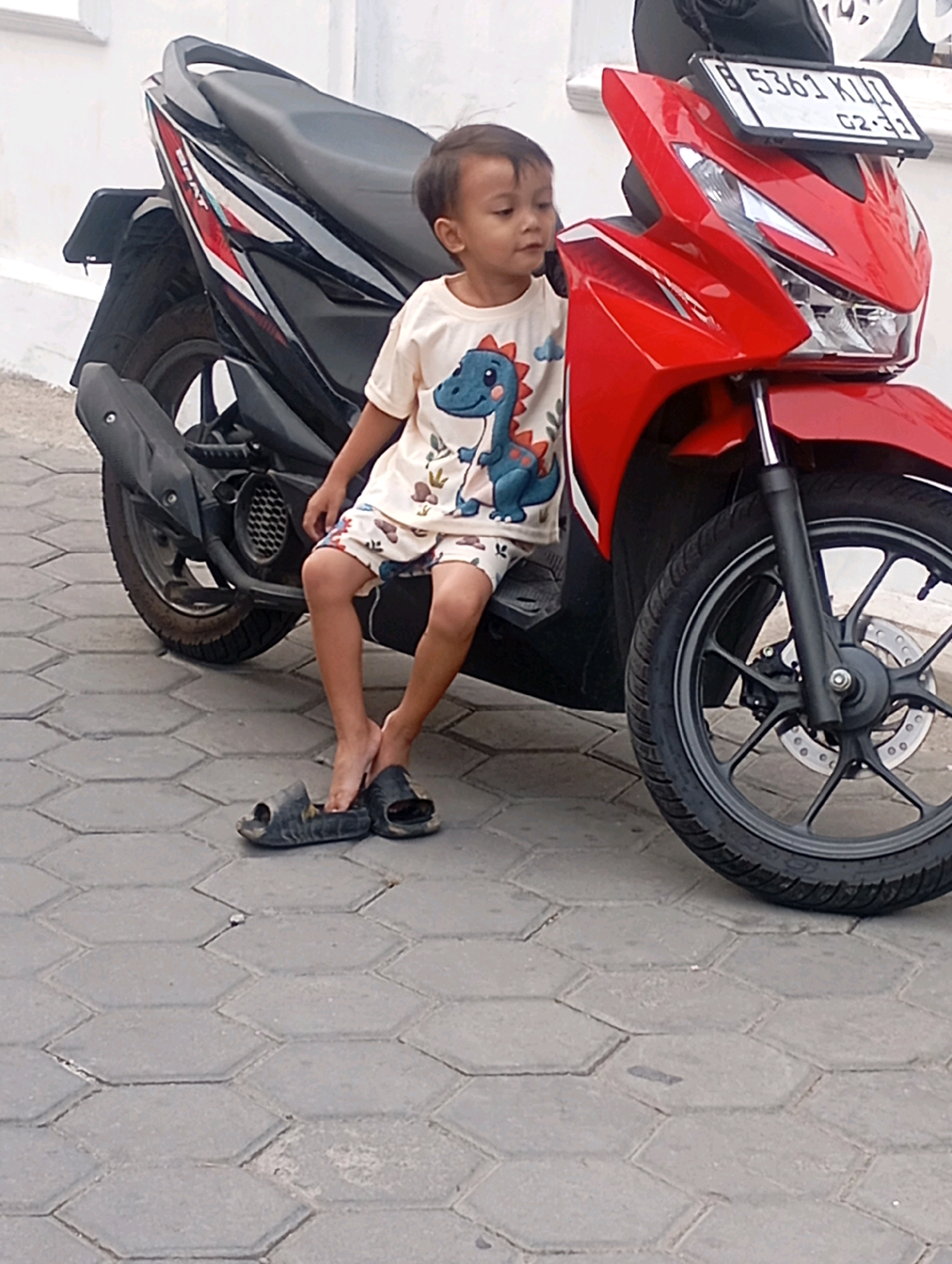 A young boy is sitting on the seat of a red and black motorcycle in Babelan, Indonesia. He is wearing a light-colored t-shirt with a dinosaur print and patterned shorts. His legs are bare, and he is wearing dark sandals on his feet. The motorcycle is parked on a paved sidewalk made of hexagonal gray tiles. In the background, a white wall is visible. The license plate on the motorcycle reads "B 5361 KLL". The overall scene appears to be an outdoor, everyday setting within the city.