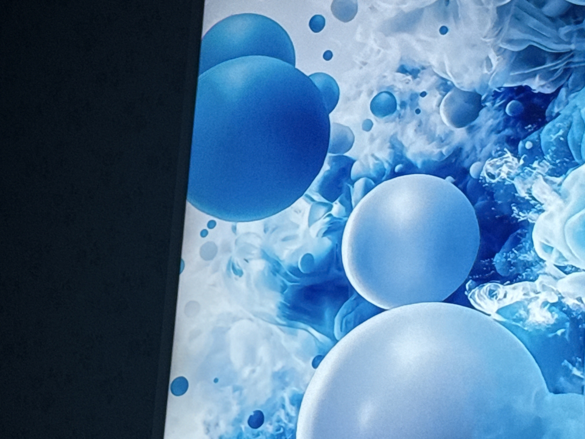 The image shows a close-up of a screen displaying an abstract design featuring blue and white spheres of varying sizes against a swirling, cloud-like background in shades of blue and white. A dark, possibly black, border occupies the left edge of the image, partially obscuring the screen's edge.
There is no information in the image to indicate its location as Mytishchi, Russia or any other location. There are no people, actions, or interactions depicted in the image.