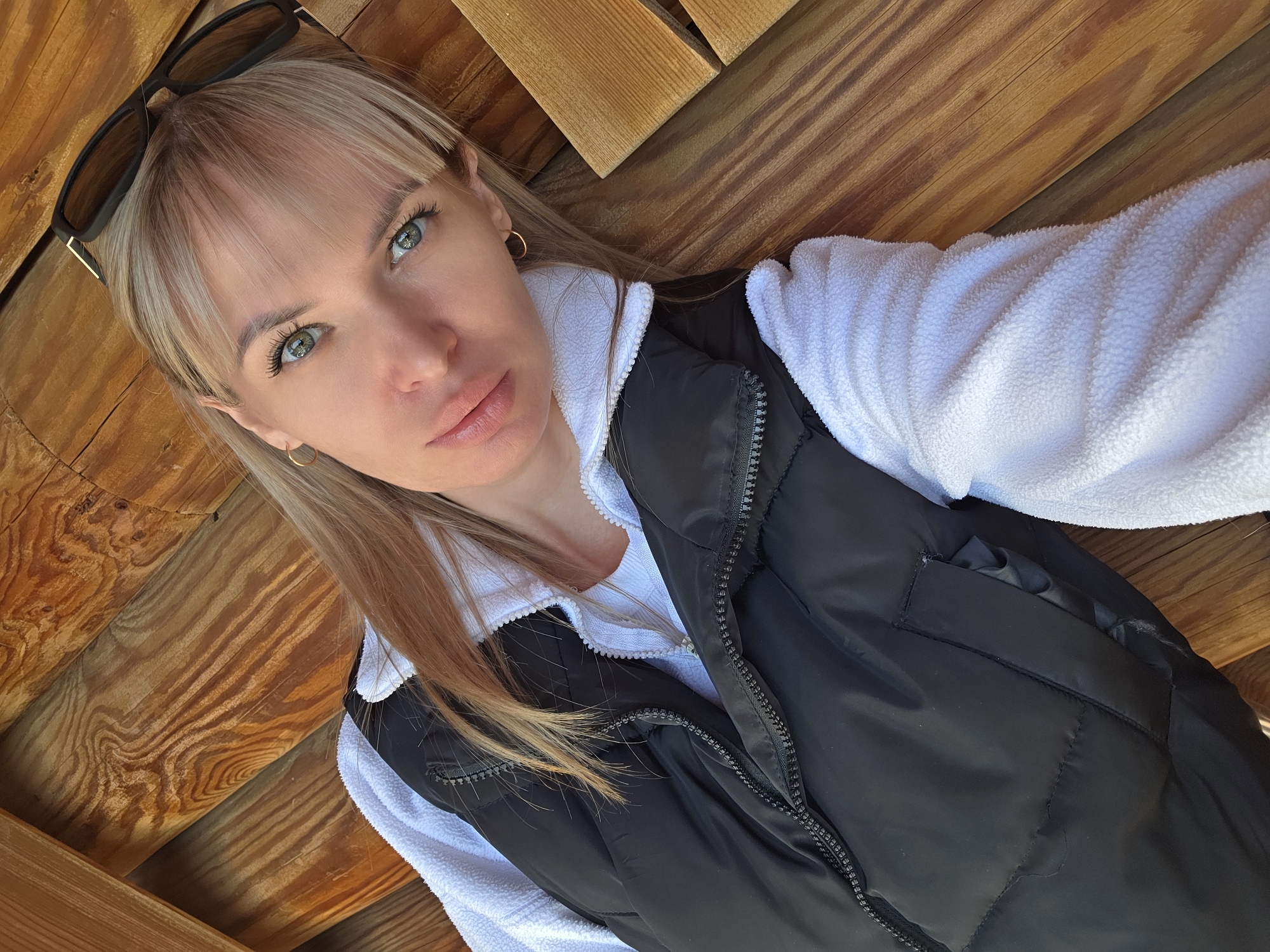 A woman is taking a selfie outdoors, laying on a wooden surface. She is fair-skinned with blonde hair and green eyes. She is wearing sunglasses on her head, small gold hoop earrings, a white collared fleece jacket, and a black puffer vest. The location appears to be a wooden structure. There is no information to determine if this image was taken in Izhevsk, Russia.
