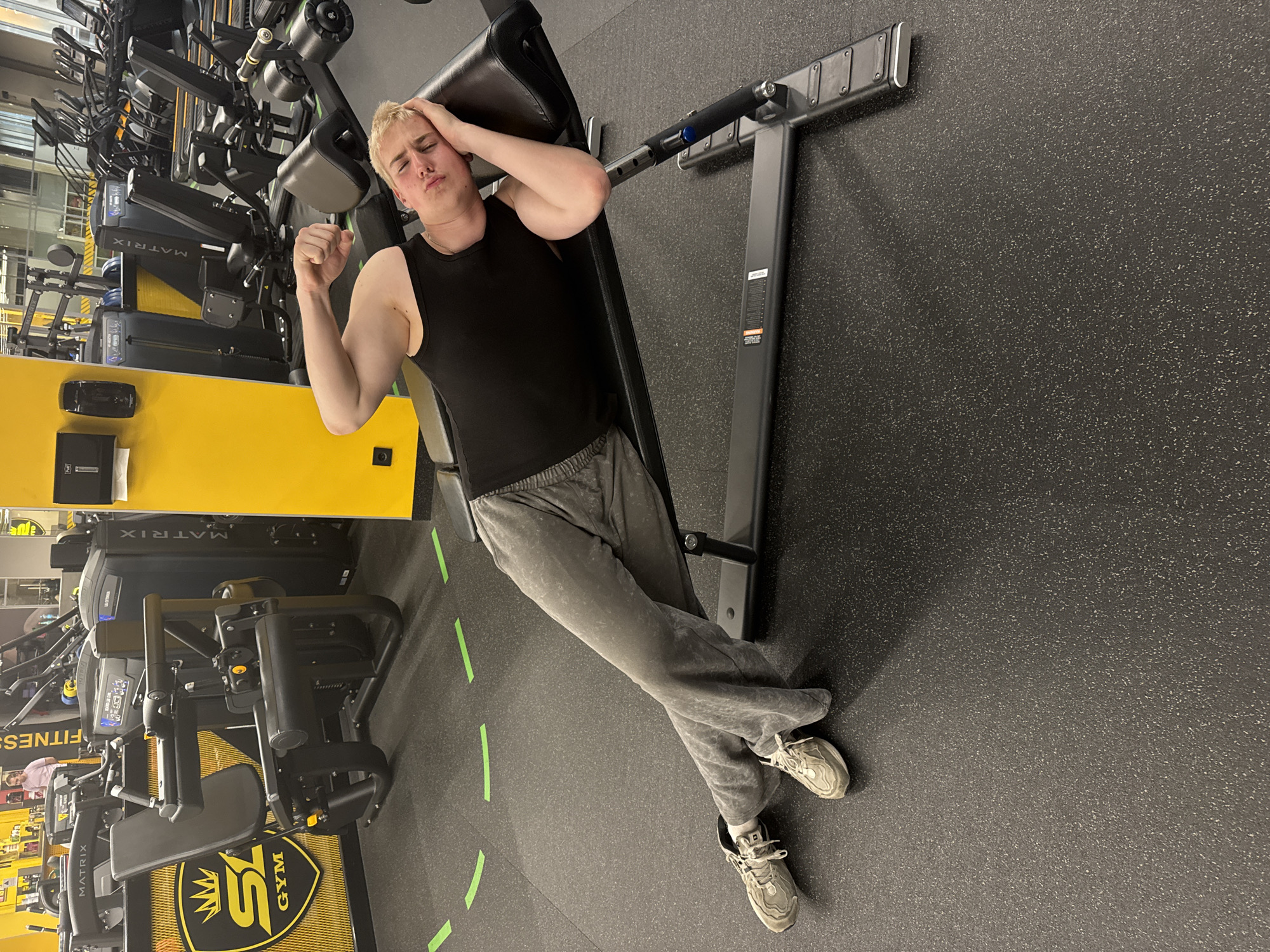 The image shows a young man lying on a black gym bench in an indoor gym environment. He has blonde hair and is dressed in a black tank top, grey sweatpants, and light-colored athletic shoes. He strikes a humorous pose, with his right hand resting on his head and his left arm flexed, while sporting a slightly pained or exaggerated expression.
The gym features a dark grey, speckled rubberized floor with green tape lines marking sections. In the background, numerous black and yellow gym machines are visible, some prominently displaying the brand "MATRIX." A yellow surface behind the man holds a black container and what appears to be a tissue box. Another piece of equipment nearby features a black and yellow shield-shaped logo with a crown and the text "SY GYM." The overall lighting suggests it is daytime. The scene conveys a playful or lighthearted moment during a workout. Additional visible text includes a partial "FITNESS" on another machine.