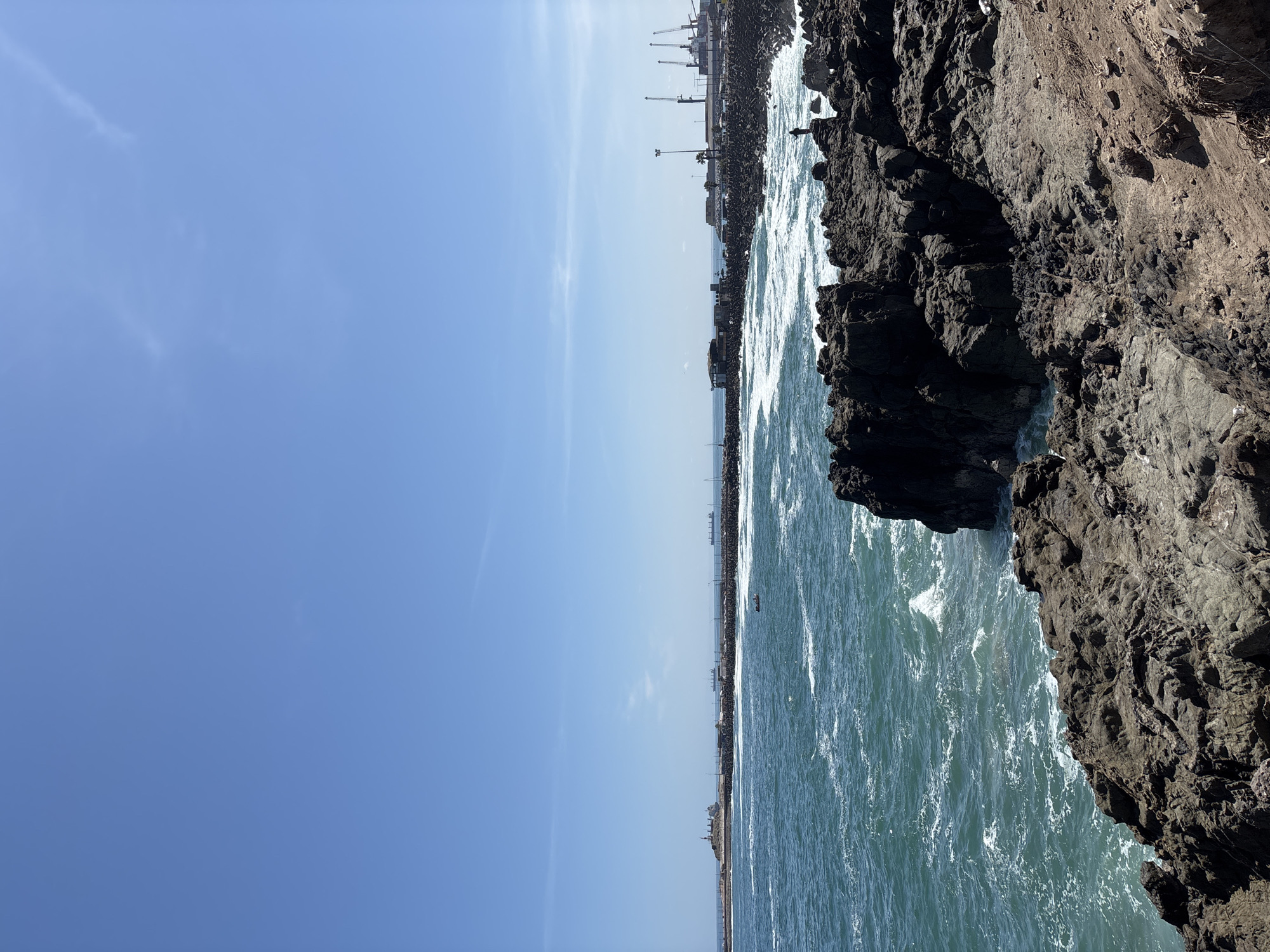 This image presents a picturesque coastal landscape in Arica, Chile, under a brilliant blue sky. The sky, mostly clear with a few subtle, wispy clouds, occupies the upper portion of the frame, suggesting a sunny and pleasant day.
The foreground is dominated by a rugged, dark grey and brown rocky cliff or shoreline, showcasing intricate textures and crevices, which creates a strong contrast with the bright water. The ocean, appearing in shades of turquoise and deep blue, is active with white, foamy waves crashing against these rocks and rippling across the surface.
Stretching across the mid-ground is a distant horizon line, where the sea meets a low-lying, built-up area. This distant shore appears to be an industrial port or harbor, characterized by several prominent, tall cranes, various buildings, and what looks like a long breakwater or artificial rock wall protecting the harbor from the open sea. Waves are also visible breaking against this distant barrier. A small, dark boat can be seen on the water in the middle distance, closer to the port structures. A tiny, dark human figure is discernible standing on the distant breakwater, seemingly engaged in an activity like fishing or observation.
The bright daylight illuminates the scene, casting distinct shadows on the rocky formations and highlighting the dynamic movement of the waves. The image captures a harmonious blend of natural coastal beauty and human industrial activity. There is no visible text in the image.