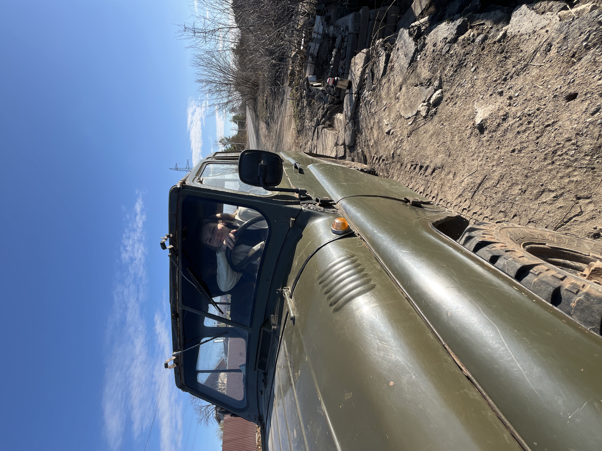 A young woman is driving an olive-green UAZ-452 van along a dirt road in what appears to be a rural area in or near Chaikovsky, Russia. The sky is bright blue with scattered clouds. The road is unpaved, with dirt and rocks. There are barren trees and some discarded materials along the roadside. The UAZ-452 is an old, utilitarian vehicle, with visible wear and tear. The location is confirmed to be in or near the city of Chaikovsky, Russia. The photograph is taken at a slightly low angle, capturing the front and side of the van, as well as the driver.