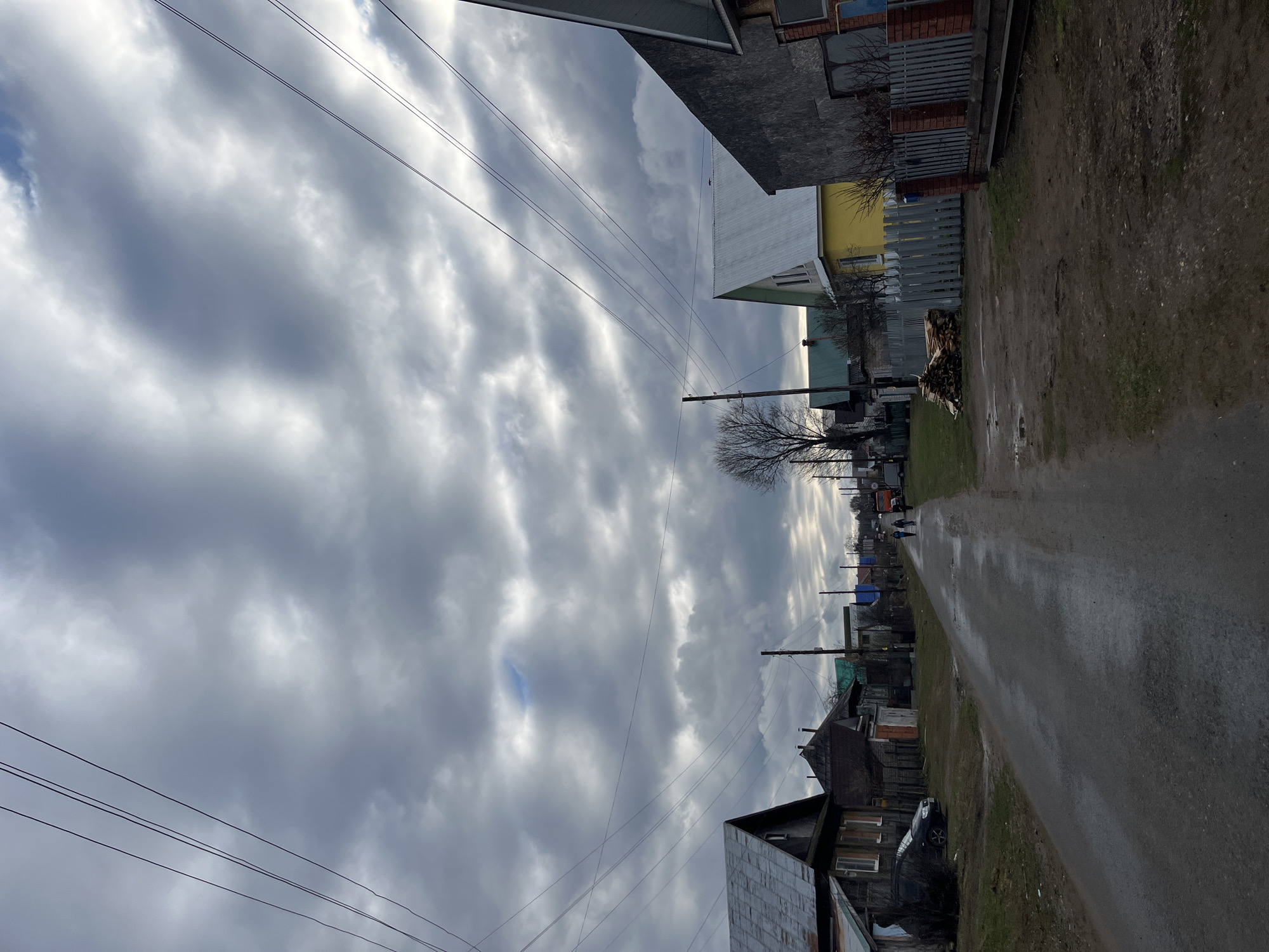 The image captures a rural or semi-rural street scene in Chaikovsky, Russia, under a predominantly overcast sky. The view looks down a narrow, unpaved or dirt road that appears damp, suggesting recent precipitation like rain or melted snow.
On both sides of the street, various single-story or 1.5-story houses are visible, featuring different architectural styles, building materials, and roof colors. Some homes have painted walls, while others show brickwork or metal siding. Fences, including wooden and metal designs, separate the properties from the street.
The sky is heavily laden with grey clouds, though some brighter patches indicate light breaking through, and a small area of blue sky is visible in the upper left. Multiple power lines traverse the sky, connected to utility poles lining the street. Bare trees, without leaves, are scattered among the houses, suggesting it is either late autumn, winter, or early spring.
Notable objects include several large piles of cut firewood stacked neatly outside a house on the right side of the road. Further down the street on the left, a dark-colored car is parked. In the distance, an orange vehicle, possibly a truck, can be seen, along with two small figures, likely people, walking down the road. The scene depicts everyday life in a residential area during a cloudy daytime. There is no visible text in the image.