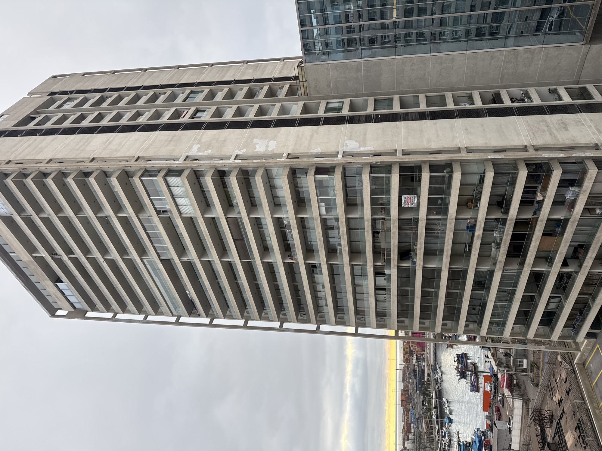 This is a high-angle, exterior, daytime shot of a modern, multi-story apartment building in Iquique, Chile. The building is constructed of concrete and features numerous balconies with glass railings, and many windows. The facade shows some weathering and discolored patches. To the right of the main building, a contemporary glass-faced building reflects the sky and surrounding structures.
In the foreground, a section of a paved surface with yellow parking lines is visible. The background reveals a bustling harbor with numerous boats docked in the water, including fishing vessels and smaller yachts. Beyond the harbor, a city skyline with industrial elements, including shipping containers, is apparent. The sky is overcast with a band of bright, warm light on the horizon, suggesting either sunrise or sunset over the ocean.
There are no discernible people in the image. The apartment building appears to be inhabited, with some balconies showing signs of occupancy, such as laundry hanging out to dry and items placed on them. A red and white "SE VENDE" (For Sale) banner is visible on one of the lower balconies of the concrete building. The overall scene depicts an urban coastal environment with a mix of residential and industrial activity.