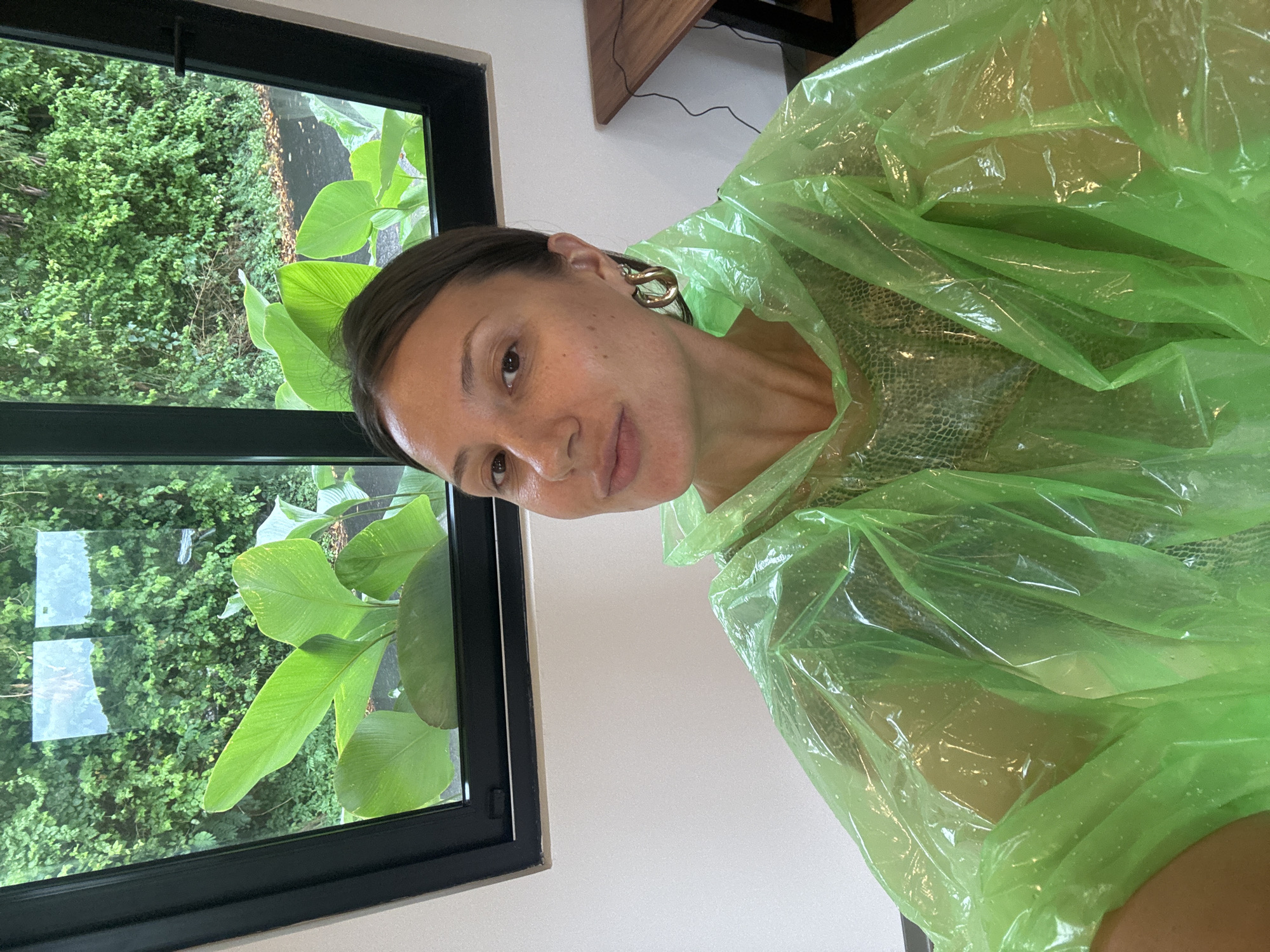 A woman is wearing a green, translucent plastic poncho, potentially for protection from rain or splashes. She is positioned indoors, with a window showing lush greenery outside. The window frame is black. The scene is brightly lit by natural light. The specific location within Kuta Selatan, Indonesia, cannot be determined from the image.