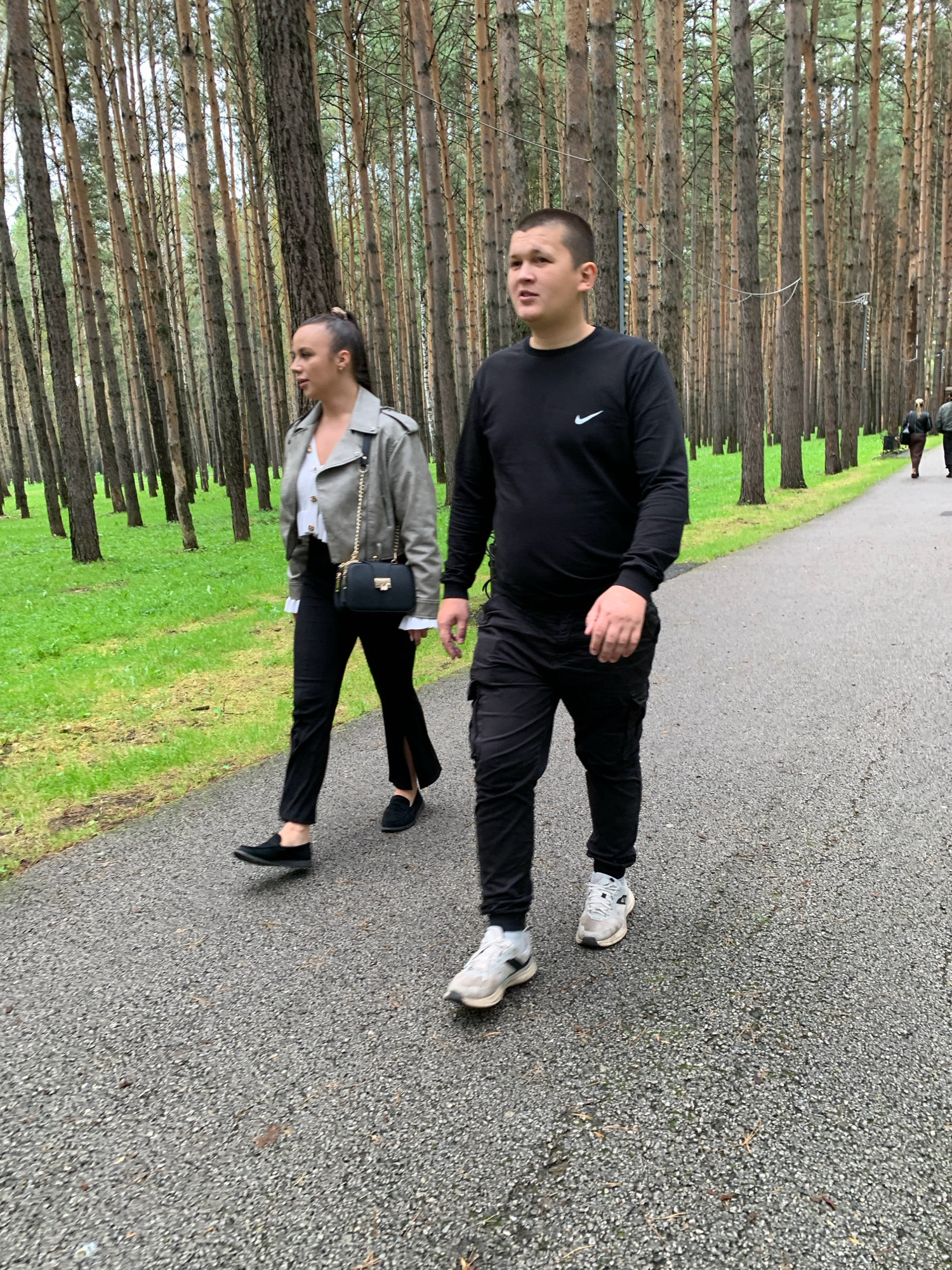 An outdoor, eye-level, medium shot shows a young man and woman walking away from the camera on a paved path through a pine forest. The path is gray and appears wet. The forest is dense with tall, slender pine trees, and there is green grass on either side of the path.
The woman, on the left, is walking with her head turned to her left. She is wearing a gray trench coat over a white shirt with ruffles, black flared pants, and black loafers. She has a black purse with a gold chain strap slung over her shoulder. Her hair is pulled back in a ponytail.
The man, on the right, is walking in profile, looking ahead. He has a buzz cut hairstyle. He is wearing a black long-sleeved shirt with a white Nike logo on the chest, black cargo pants, and white athletic shoes.
In the background, further down the path, two other figures are visible, walking in the same direction. The lighting suggests an overcast day. The location is identified as Prokopyevsk, Russia.