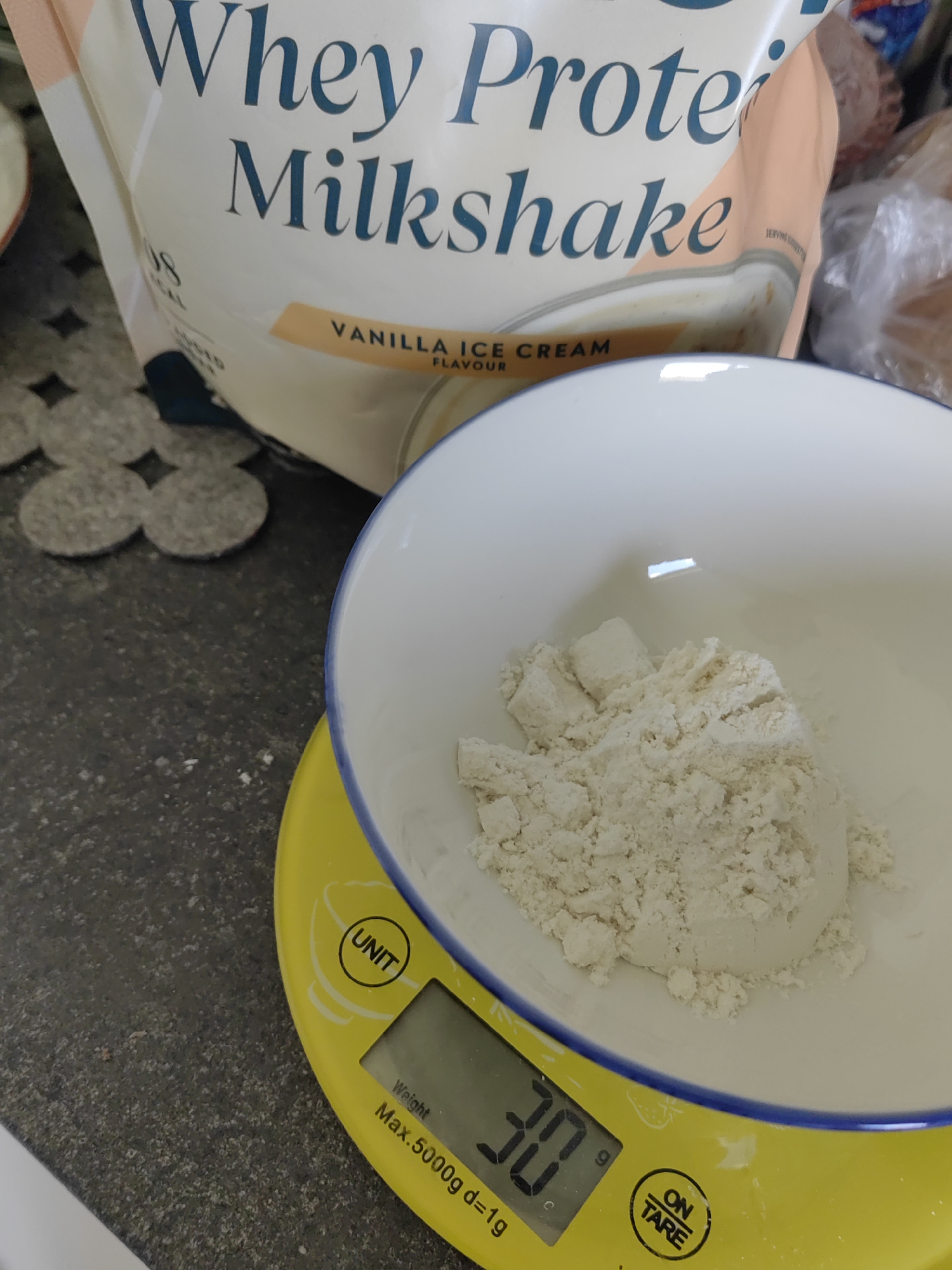 The image displays a close-up, overhead view of a kitchen counter, likely in a home. The background features a large bag of "Whey Protein Milkshake" with "Vanilla Ice Cream flavour" clearly visible. In the foreground, a white bowl containing a light-colored powder, presumably protein powder, rests on a yellow digital scale. The scale displays a weight of "30g". The surrounding counter surface is dark and speckled, and a portion of a white and blue patterned bowl is also visible. The scene suggests the process of measuring protein powder for a drink or meal. There are no people or animals present in the image, and the environment appears to be indoors. There is no indication of the city of Djerovo, Bulgaria.