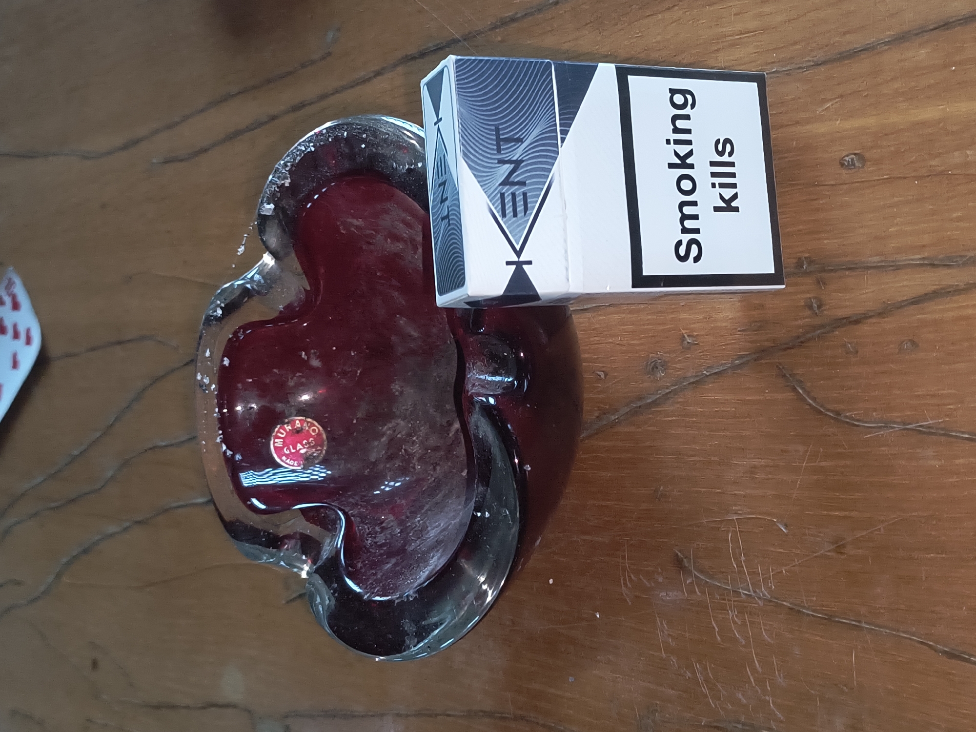 A pack of Kent cigarettes and a red glass ashtray sit on a wooden table. The ashtray has a sticker that reads "Murano Glass". The cigarette pack has the warning "Smoking kills" printed on it. The image is a close-up shot, and the background is blurred. There are no people visible in the image. The image does not provide any information about the location in the city of Rivne, Ukraine.