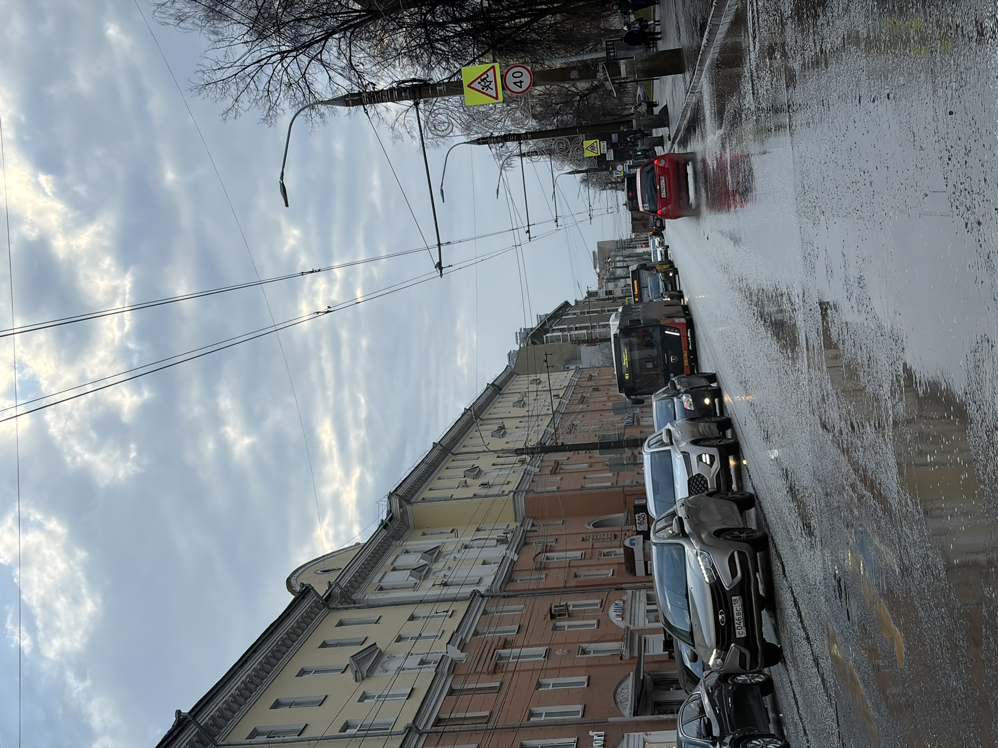 The image shows a street scene in Izhevsk, Russia, on a wet day. Buildings line the left side of the street, varying in color from light yellow to brown with multiple windows and architectural details. Overhead power lines stretch across the sky. Cars are parked along the right side of the street, and a bus and other vehicles are visible further down the street. The road surface is wet and reflects the sky. A traffic sign indicating a pedestrian crossing and a speed limit of 40 km/h is visible near the center of the frame. The sky is overcast with patterned clouds.