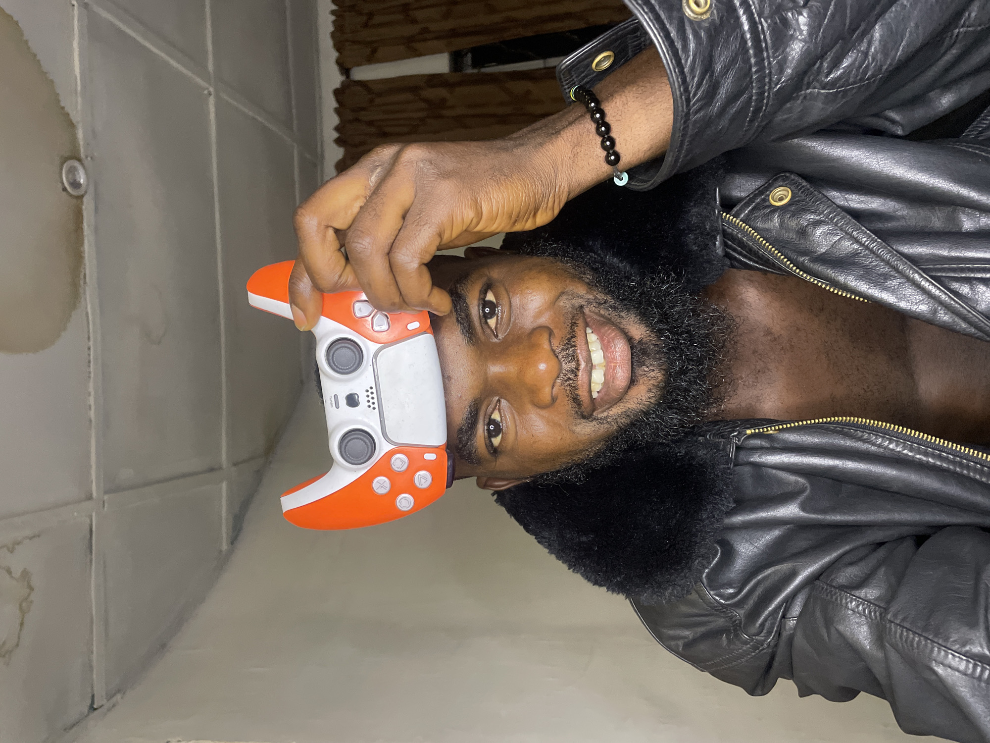 An adult man with dark skin is holding a video game controller to his head and smiling. He has a full beard and wears a black leather jacket with a fur collar and a beaded bracelet on his left wrist. The controller is white and orange. The background appears to be a tiled wall with some discoloration. The location is possibly in Jos, Nigeria.