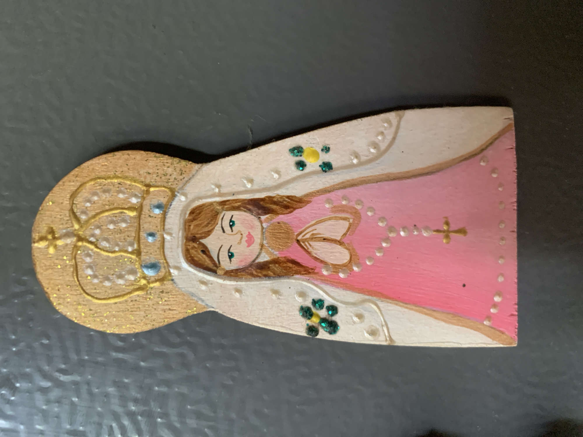 This media file captures a close-up of a hand-painted wooden decorative object, likely a magnet, depicting a stylized female figure against a dark, textured background. The object is vertically oriented as if standing upright.
The central subject is a serene female figure with fair skin, brown hair, green eyes, and pink lips. She is adorned with a golden crown, embellished with outlines and small light blue accents, suggesting jewels. Her attire consists of a bright pink garment, possibly a dress or robe, and a white cloak or mantle that drapes over her shoulders and arms. The white cloak is decorated with small, white raised dots along its edges, and two distinct green glittery floral or clover-like embellishments, one on each side. The pink garment also features a pattern of small white dots.
The figure's left hand (from her perspective) is shown holding a beaded chain, which strongly resembles a rosary, ending in a small golden cross, resting against her pink garment. Her right hand (from her perspective) is gently curved near her chest. The painting style is naive or folk art, characterized by its simplicity and bright colors. Gold glitter is visible on the crown and parts of the white cloak.
The background is a dark, likely black or charcoal gray, flat surface, suggesting the item is displayed on a refrigerator or similar magnetic surface. There is no visible activity, as it is a static image of an object. No text is visible on the object or in the background. Given the location context of Santa Ana, Venezuela, the figure may represent a local religious icon or a depiction of the Virgin Mary, possibly Our Lady of Santa Ana.