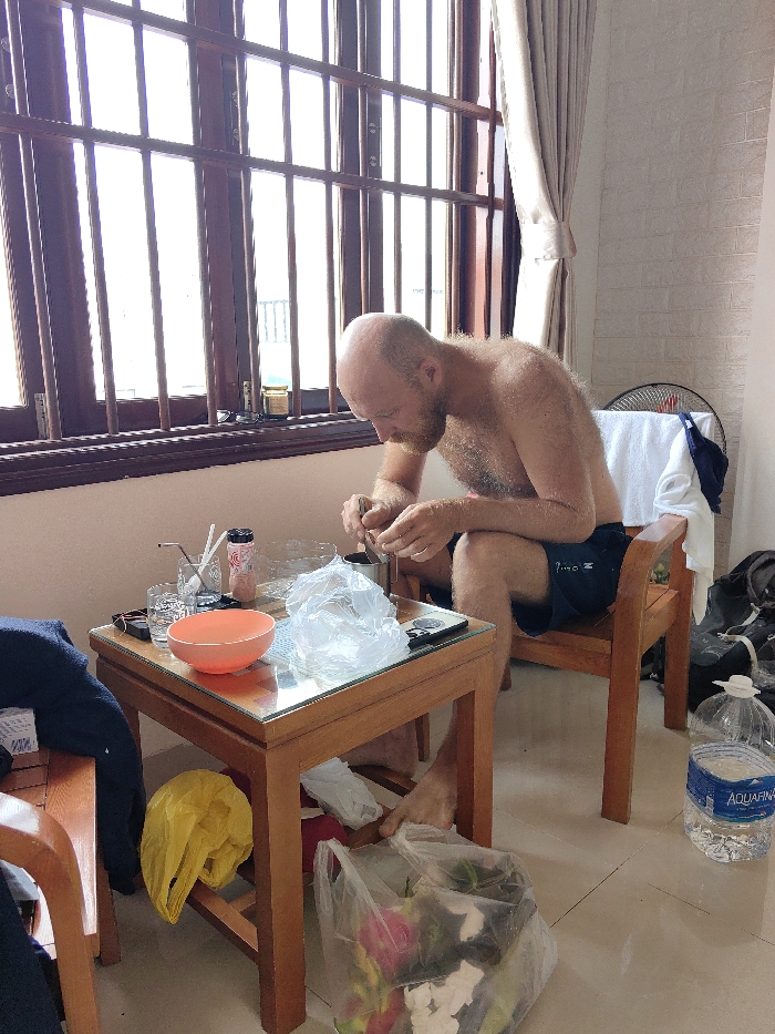 An indoor scene in Thuận Hóa Ward, Vietnam, depicts a barefoot Caucasian male, bald with a beard, shirtless, and wearing dark shorts, seated on a wooden chair. He is positioned at the right of the frame, facing left, and actively engaged in manipulating contents within a dark metal container on a small, light brown wooden table with a glass top. The table holds an orange-red bowl, several clear glasses with straws, a pink bottle, a crumpled clear plastic bag, and a dark electronic device. A large window with dark wooden frames and vertical security bars occupies the background, admitting bright natural light. Light beige curtains are drawn to the right of the window. Behind the seated male, a white oscillating fan is visible, draped with a white towel. The floor is light-colored tile. Beneath the table, a yellow plastic bag hangs. To the right of the table, on the floor, a large clear plastic bag contains fruit waste, and two clear plastic water bottles, one an Aquafina brand, are present.
