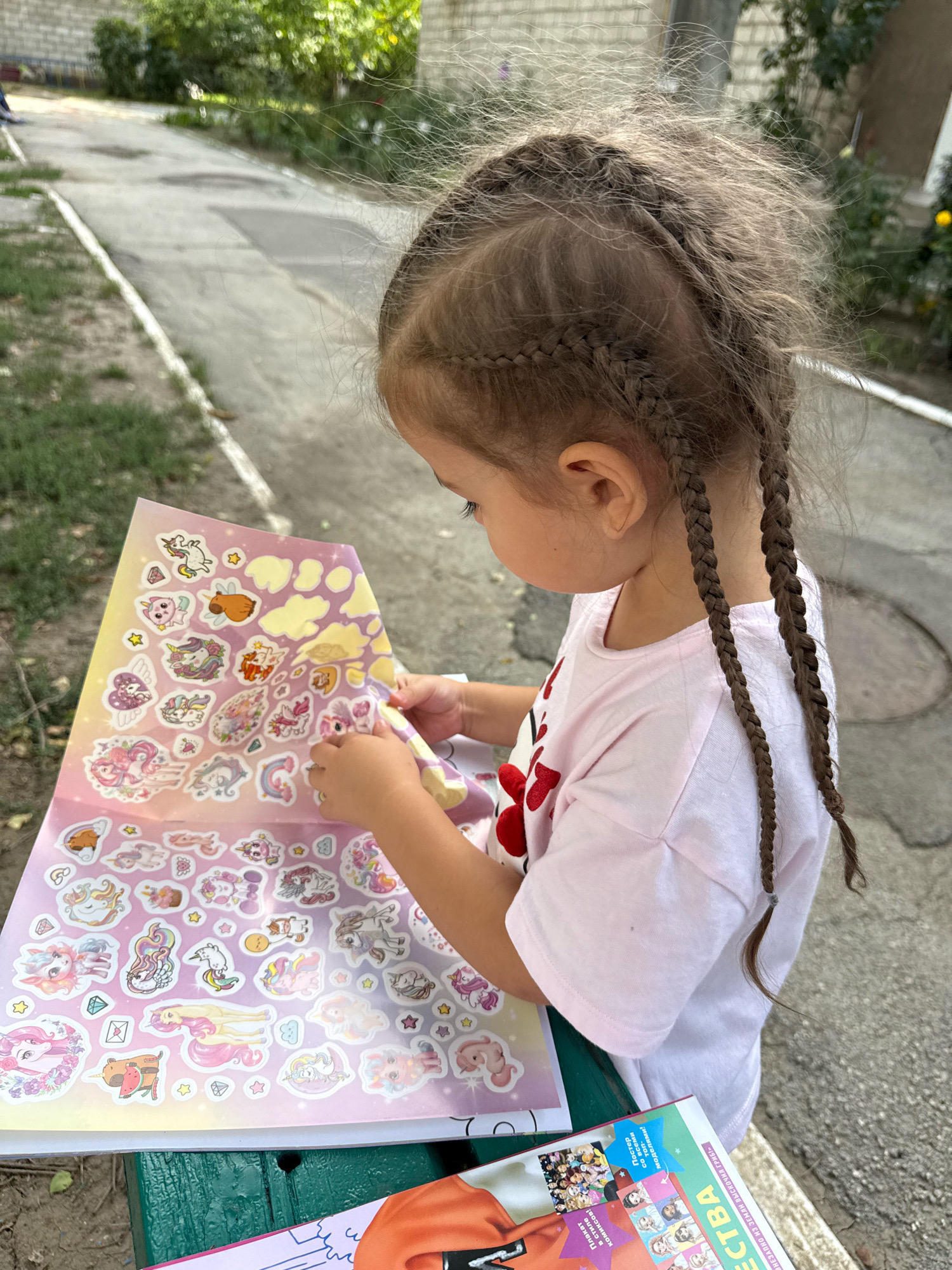 A young girl with braided hair is sitting outdoors, engaged in applying stickers from a large sheet adorned with unicorns and clouds. She is wearing a light pink t-shirt. The scene appears to be in a residential area, possibly a courtyard, with a concrete pathway, greenery, and a brick wall visible in the background. A portion of a book with colorful images is also visible on the bench where the girl is seated.