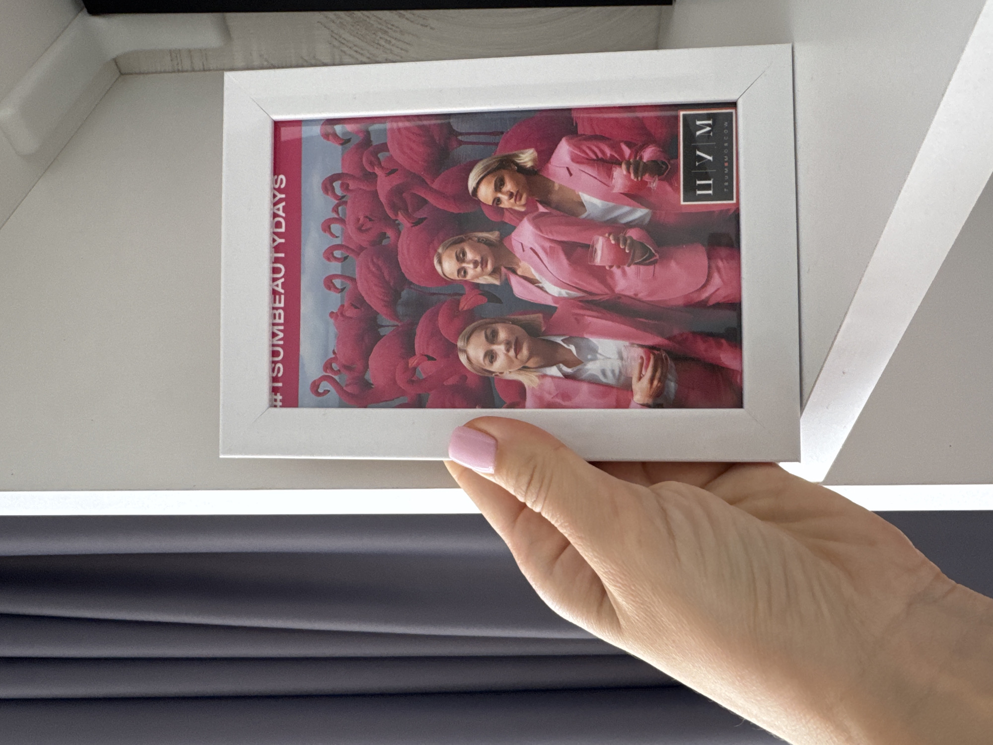 A close-up shot captures a hand holding a framed photograph. The photograph inside the frame displays three women in pink suits, each holding a drink. They are superimposed over an image of flamingos. The photograph also contains text in a box that reads "TSUM MOSCOW", and hashtags "#TSUMBEAUTYDAYS" in a strip along the edge. The frame appears to be white.
The background shows a gray, possibly draped, curtain-like fabric in the lower portion of the frame. The upper portion reveals a white surface that could be a wall or ceiling, adding to the indoor setting of the image.
The photograph is likely being held indoors. The photograph shows women associated with the shopping mall TSUM in Moscow, Russia. The image does not include Sochi, Russia.