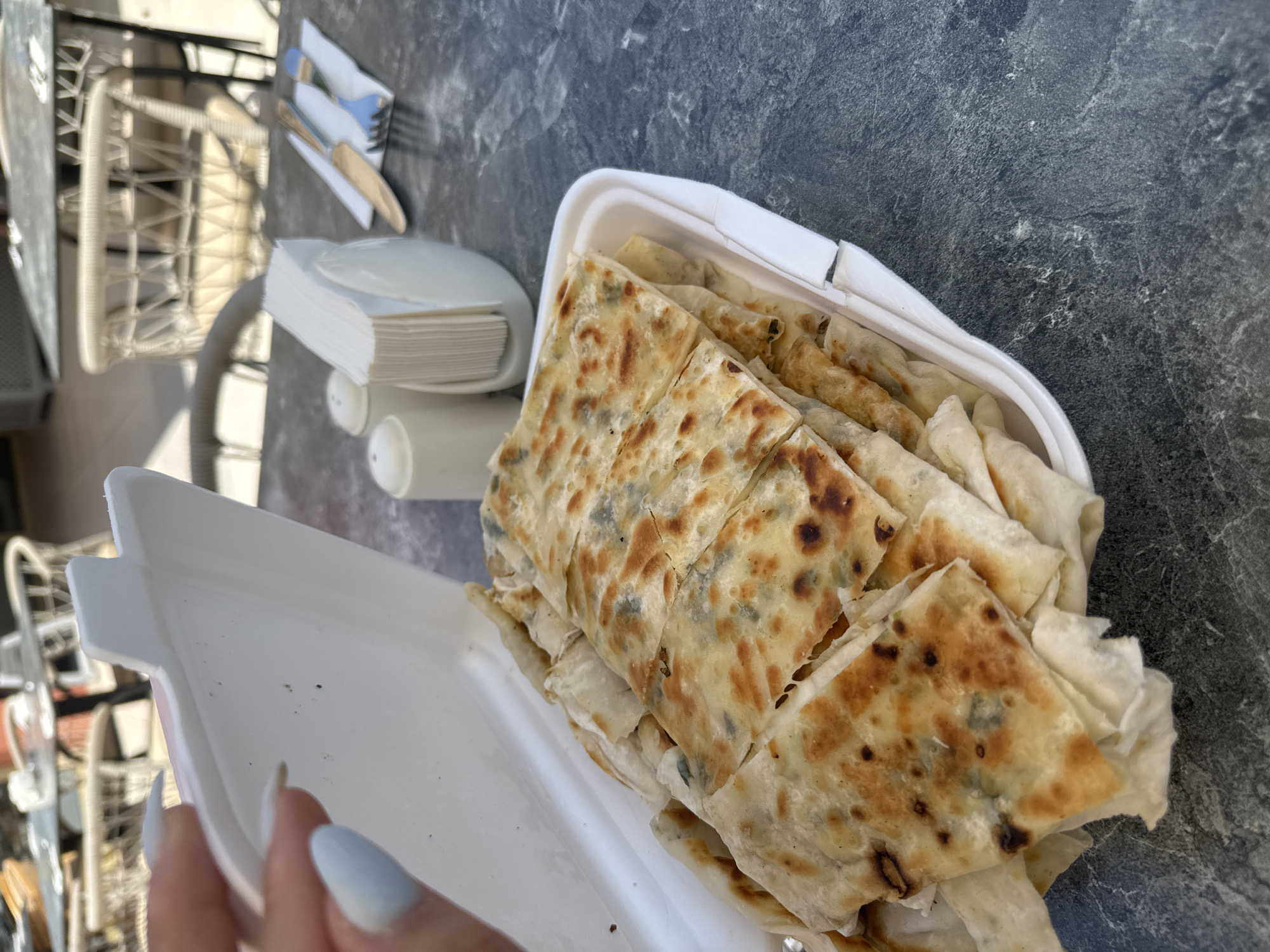 A person's hand with light blue nail polish holds the edge of a white takeout container filled with folded, grilled flatbreads. The flatbreads, appearing golden brown with darker charred spots and visible green filling, are stacked in the container. On a dark, textured tabletop behind the container are a white napkin dispenser filled with folded napkins, two white condiment shakers, a knife with a gold handle, and a blue fork. In the background, blurred outdoor seating with white woven chairs and tables is visible, suggesting a restaurant or cafe setting in Kemer, Turkey.