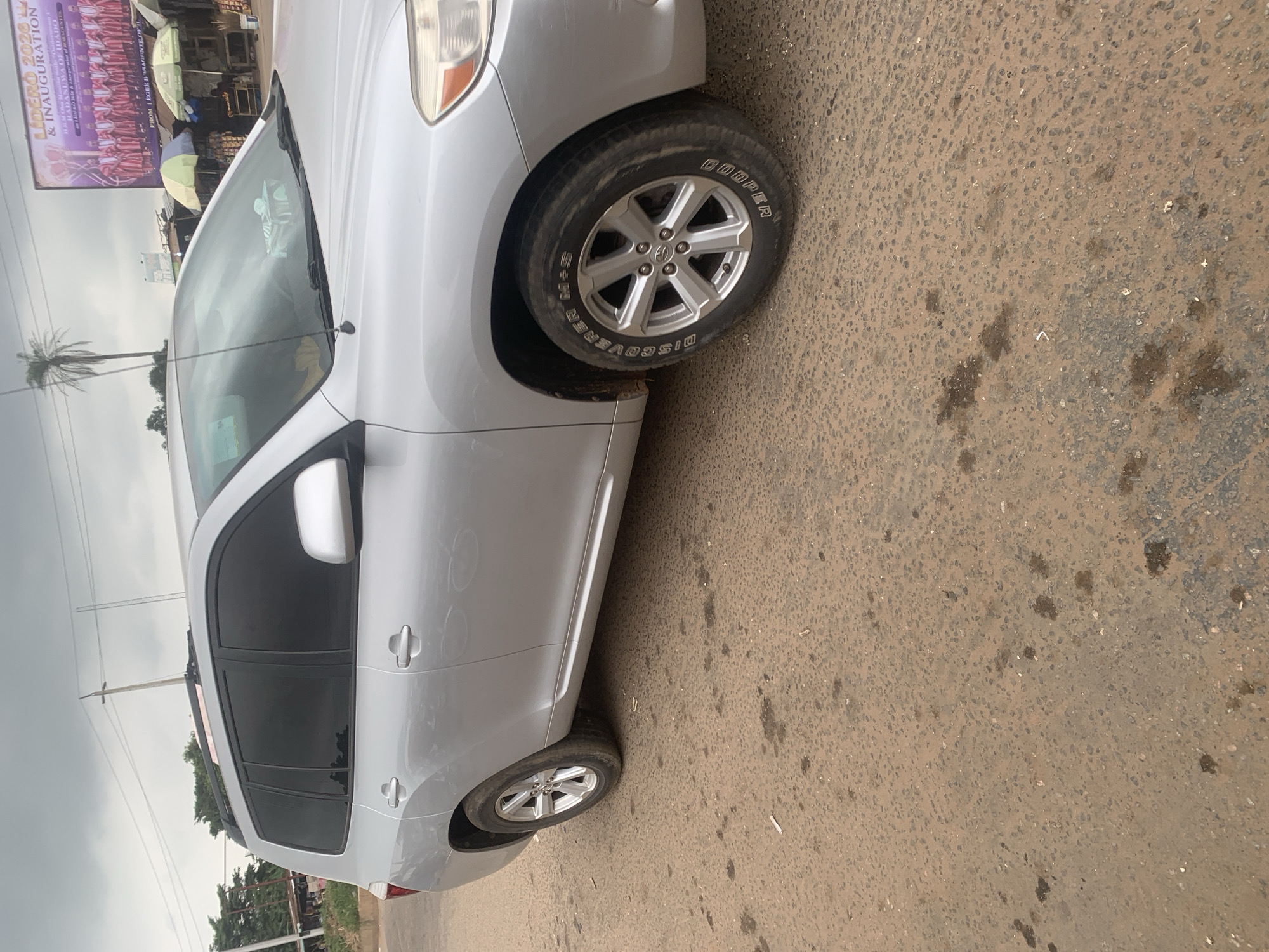 The image is a medium shot of a silver SUV parked on a dusty, paved road in Shagamu, Nigeria. The SUV is a Toyota Highlander, and its right side is prominently featured. The vehicle has tinted windows, a roof rack, and silver alloy wheels. The tires are Cooper Discoverer M+S. In the background, there's a billboard advertising a political event, "LIDERO 2026 & Inauguration," with images of people in robes. There are also market stalls with umbrellas and some greenery, including a palm tree. The sky is overcast. The road surface is rough and appears to be mixed with dirt and gravel, with some dark spots that could be oil stains. There are no people visible in the immediate vicinity of the car.