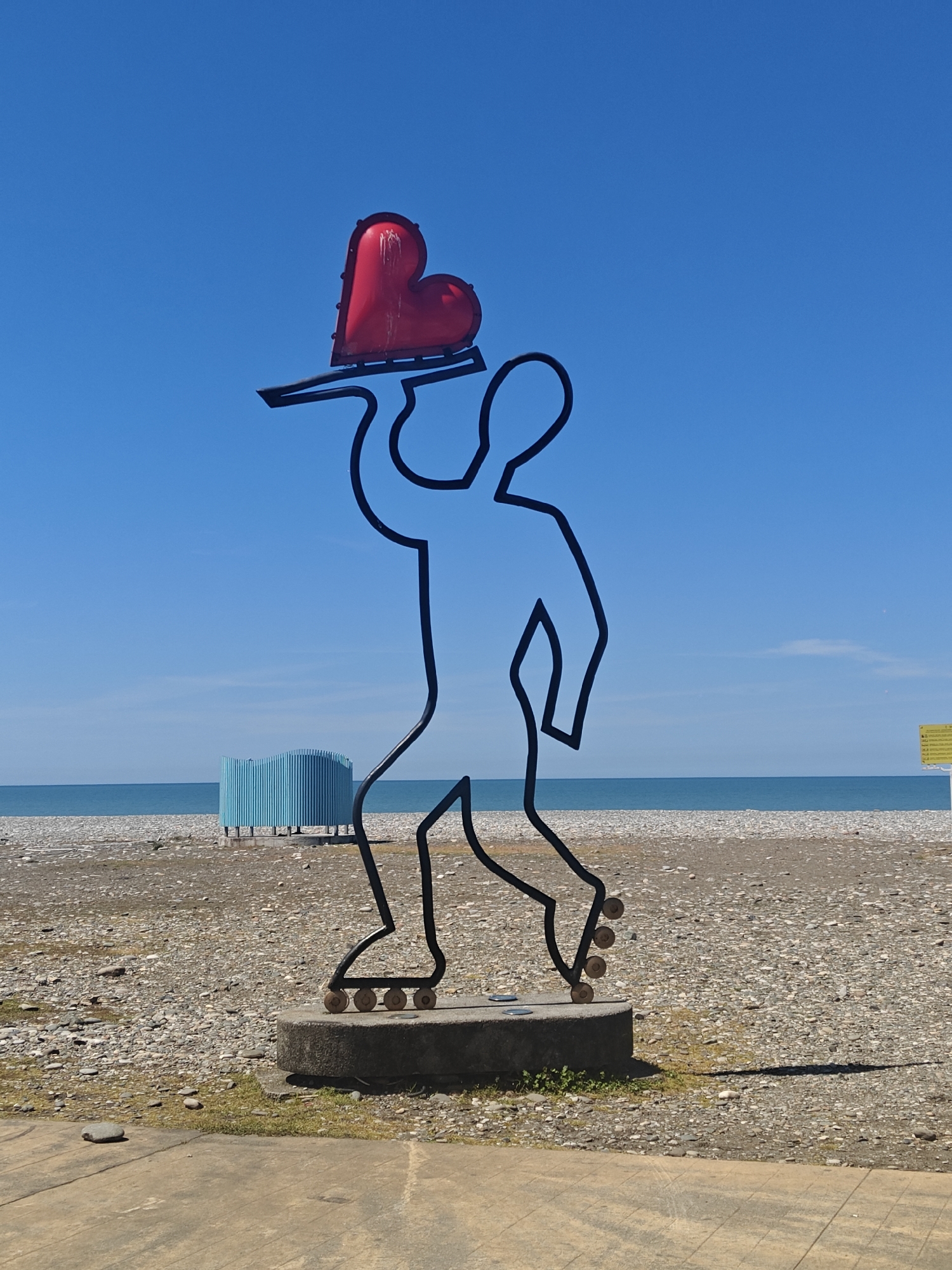 The image shows a metal sculpture of a person on roller skates holding a red heart on a tray above their head. The sculpture is located on the beach in Batumi, Georgia. The beach is made of pebbles, and the sky is blue. A blue structure stands on the beach in the background.