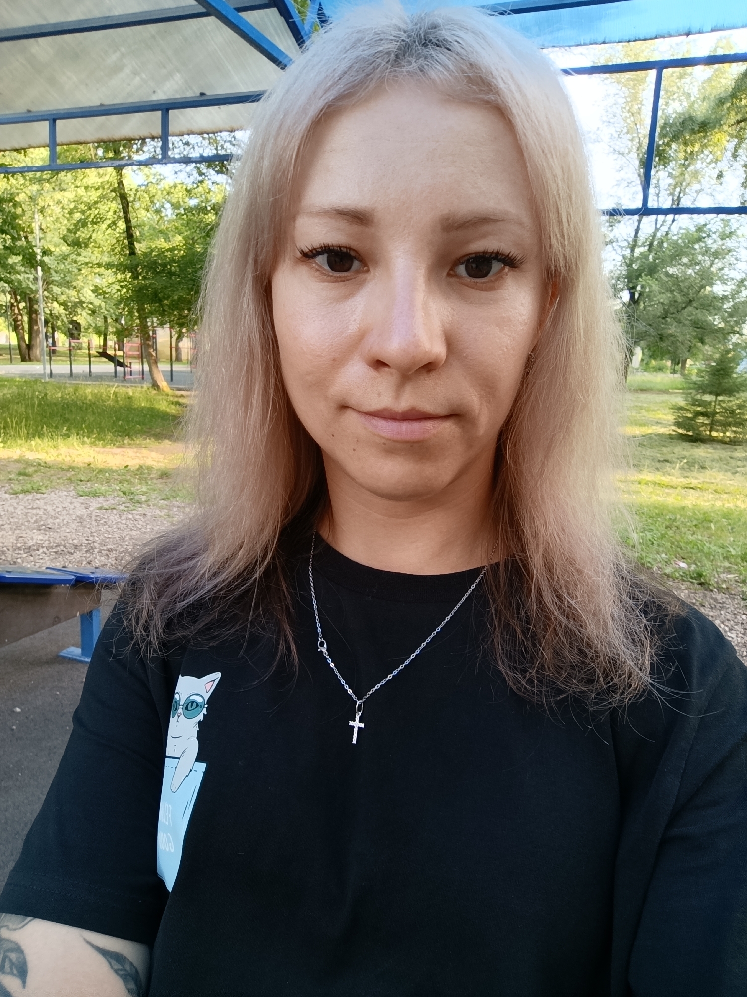 A young Caucasian woman with blonde hair pulled back from her face is posing for a selfie outdoors. She is wearing a black t-shirt with a graphic of a cat in a pocket and a silver necklace with a cross pendant. The background shows a park or recreational area with trees, grass, and playground equipment. The image was taken in Novokuybyshevsk, Russia, on a sunny day.
