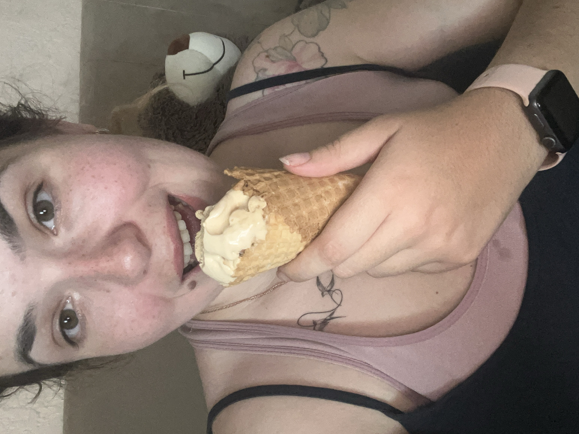 The image depicts a close-up, indoor scene. A young woman is holding an ice cream cone to her mouth, with her eyes looking towards the camera. She has freckles on her face and is wearing a pink tank top layered over a black garment. A tattoo of two birds is visible on her chest, and another floral tattoo is on her upper arm. A stuffed animal is partially visible behind her. A smartwatch with a pink band is on her wrist. The background appears to be a couch or similar furniture. The scene does not provide any specific visual cues or landmarks that would allow for the identification of its location within the city of Krasnyye Barrikady, Russia.