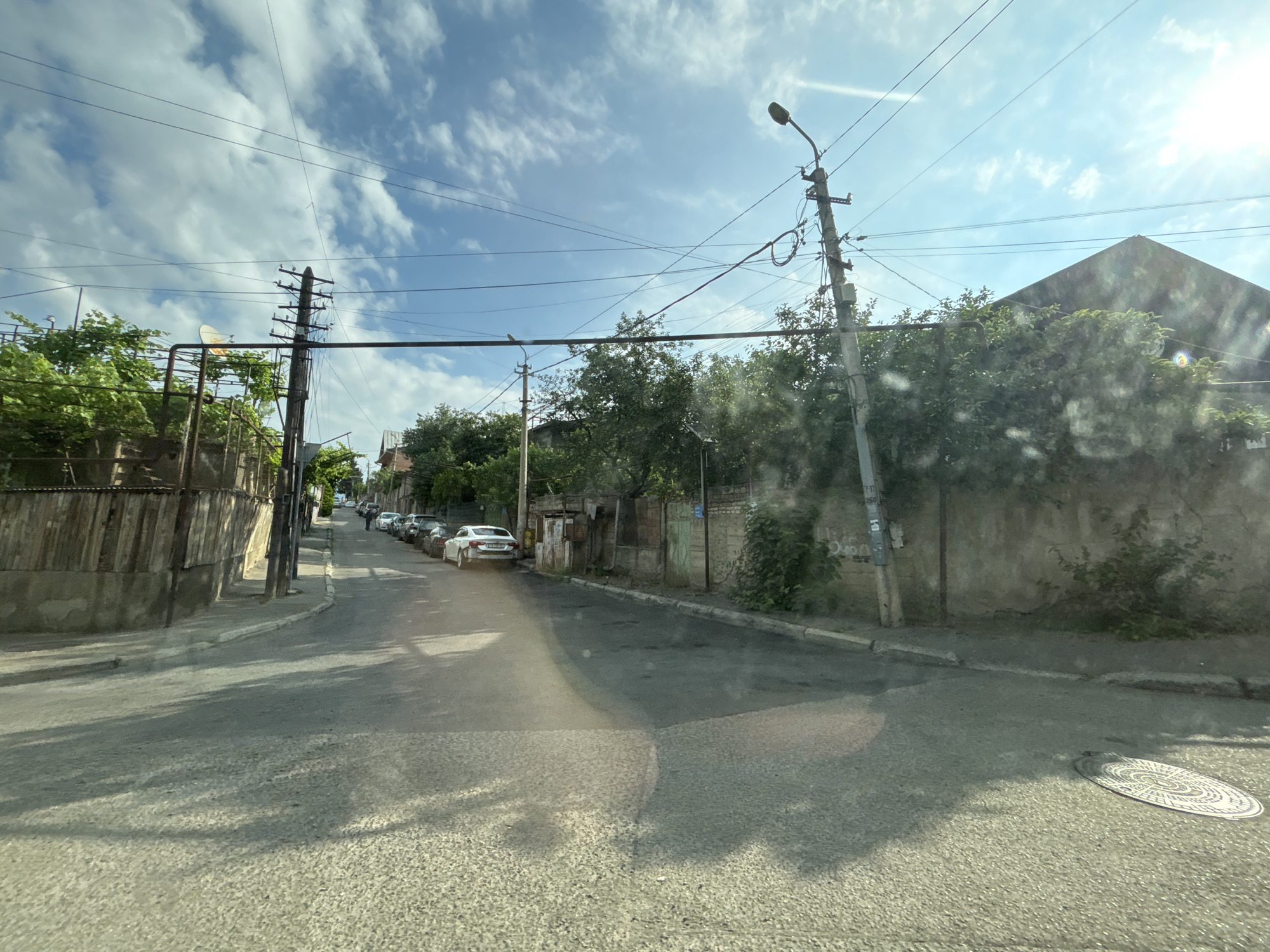 The image depicts a street scene in Tbilisi, Georgia, likely on a sunny day with scattered clouds. A paved road curves to the left, lined with older buildings and residential houses. Trees and foliage are present along the sides of the street, with some overgrown vegetation. A series of utility poles with wires crisscross the sky. Several parked cars, including a white sedan and other sedans and hatchbacks, are visible along the side of the road. A man is standing near the parked cars further down the street. A manhole cover is visible on the right side of the road. The architecture suggests a mix of older, possibly Soviet-era structures, with some newer elements. The overall impression is of a typical, somewhat aged urban residential street.