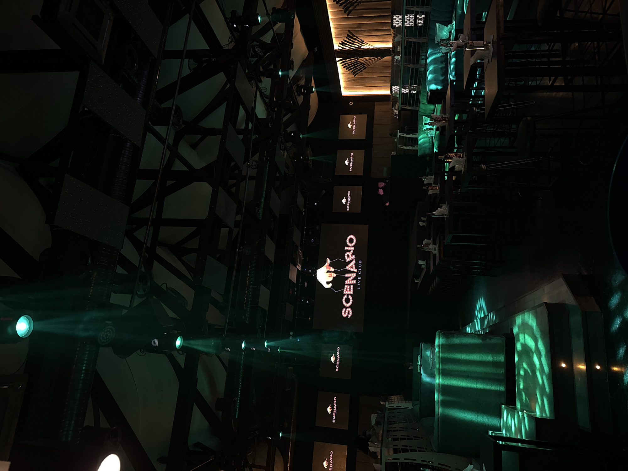 The image shows the interior of a dimly lit club called "Scenario Live Club" in Tsarevo, Bulgaria. Green lights illuminate the space, casting patterns on the floor and furniture. Several screens display the club's logo. Booth seating and tables are visible, with some bottles of alcohol on them. A DJ booth is also present. The club appears to be empty or sparsely populated. The architecture includes exposed beams and a stage area.