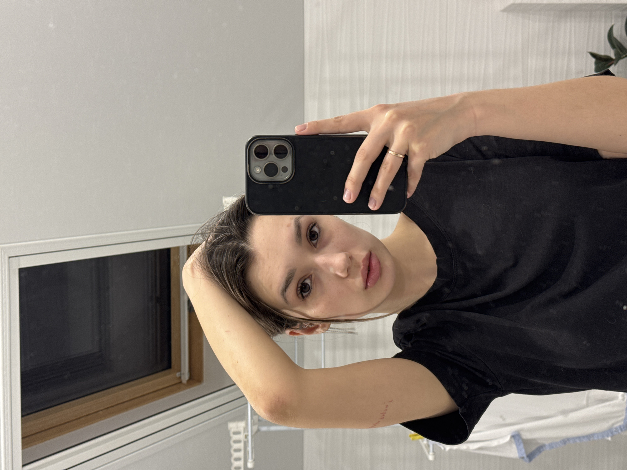 A woman is taking a selfie in a bathroom. She is holding a black smartphone with three camera lenses in her right hand, which is adorned with a simple gold ring. She wears a black t-shirt. Her brown hair is styled loosely, and she looks directly at the camera with a neutral expression.
The background reveals a typical bathroom setting. A window with a dark shade is visible behind her to the left. The walls are painted a neutral grey. To the right, a small portion of a houseplant can be seen. The location is not identifiable.