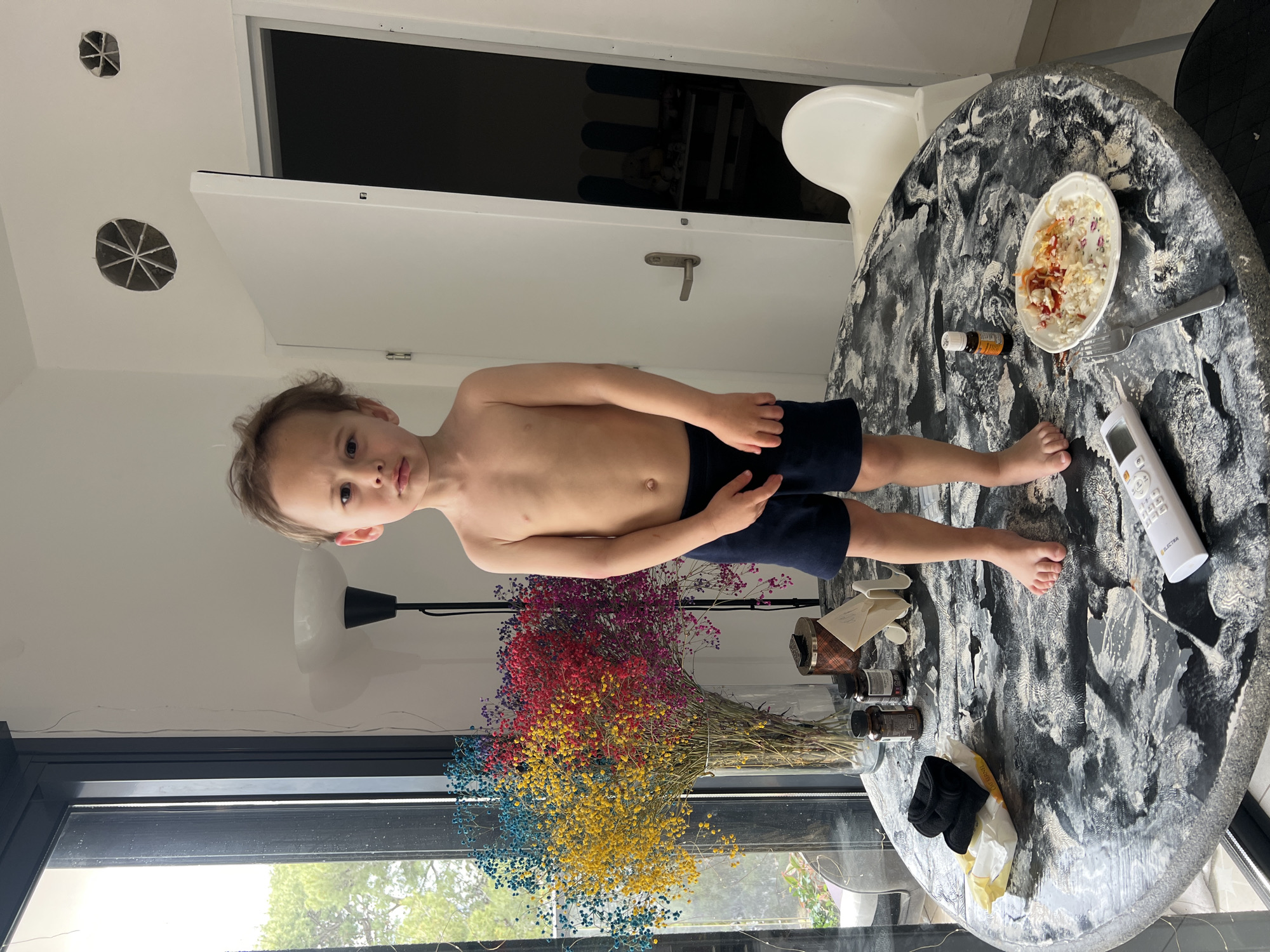 A shirtless child stands on a table covered in a white powdery substance. On the table, there is a plate of food, a remote control, and other miscellaneous items. A bouquet of colorful flowers is visible near a window. The location is Kiryat Bialik, Israel.