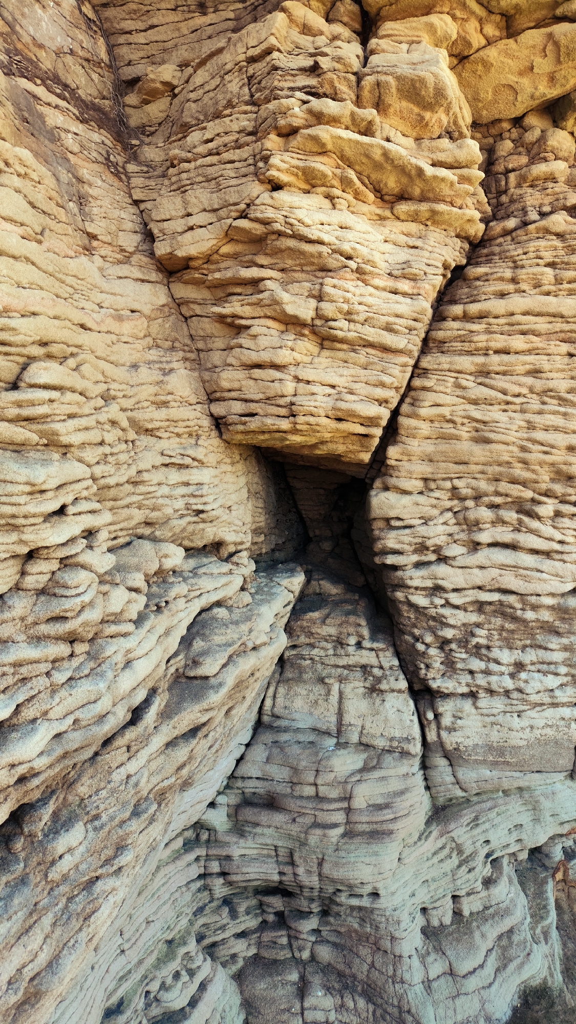 The image shows a close-up, eye-level view of the textured sandstone cliffs at Red Rocks, West Kirby, United Kingdom. The rocks are layered with horizontal striations, varying in color from light tan to darker grey-brown. A small, dark opening or crevice is visible in the center of the frame, formed by the jutting angles of the rock formations. The natural erosion has created a rugged and uneven surface across the entire scene.