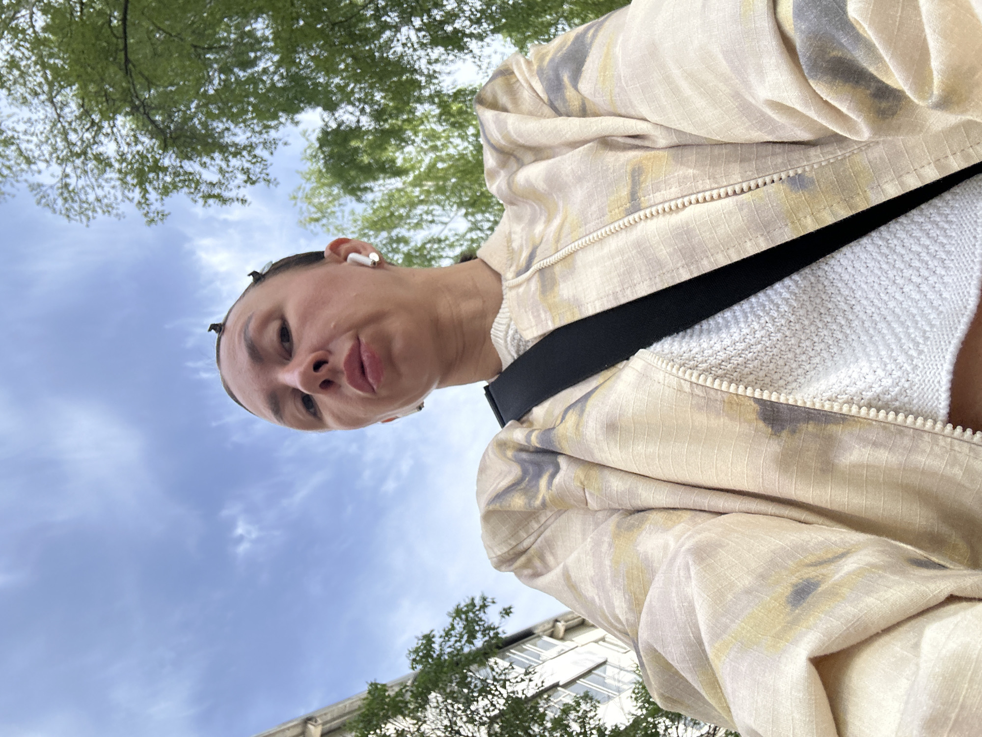 An upward-facing selfie photograph shows a person wearing a white sweater underneath a zippered, patterned tan jacket with a black strap across the chest. The person is wearing a white wireless earbud. Above the person are trees and a blue sky with light clouds. The bottom of a building can be seen at the bottom left of the image. The exact location in the city of Votkinsk, Russia cannot be determined from the image.