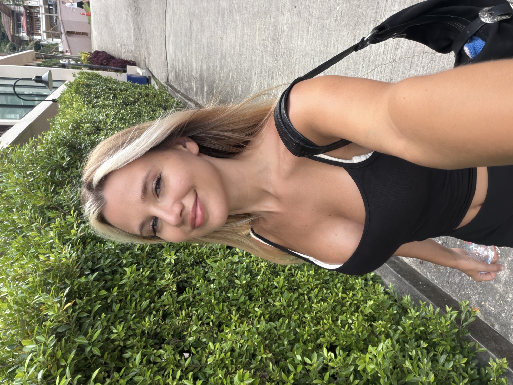 A young woman with blonde hair, wearing a black tank top and black pants, smiles at the camera while walking down a paved sidewalk next to a thick green hedge. She is holding a clear plastic water bottle in her right hand and a black purse with a silver buckle is slung over her left shoulder. The background shows a hilly street with buildings and trees. The location is identified as Wichit, Thailand.